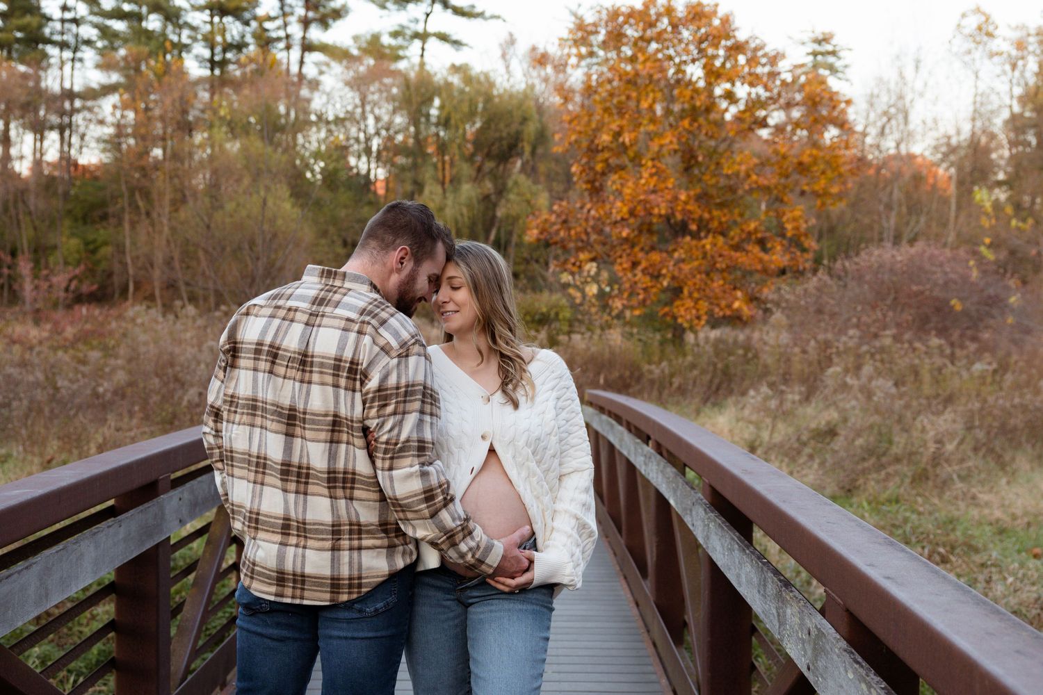 Couple holding baby bump on wooden bridge surrounded by colorful autumn foliage during maternity photo session.
