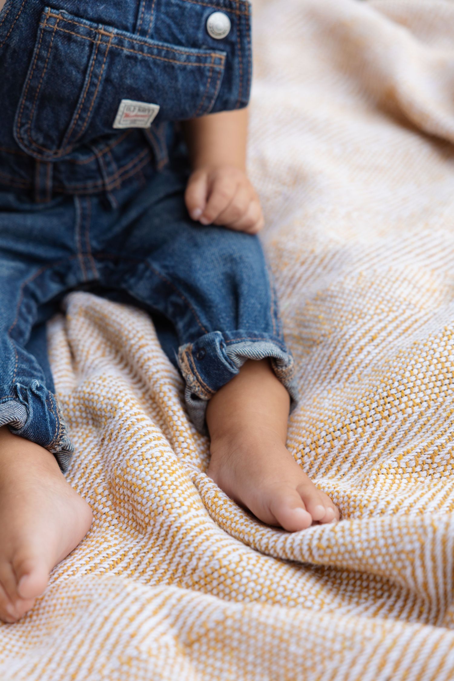 Close-up of tiny feet in denim overalls resting on a textured cream blanket.