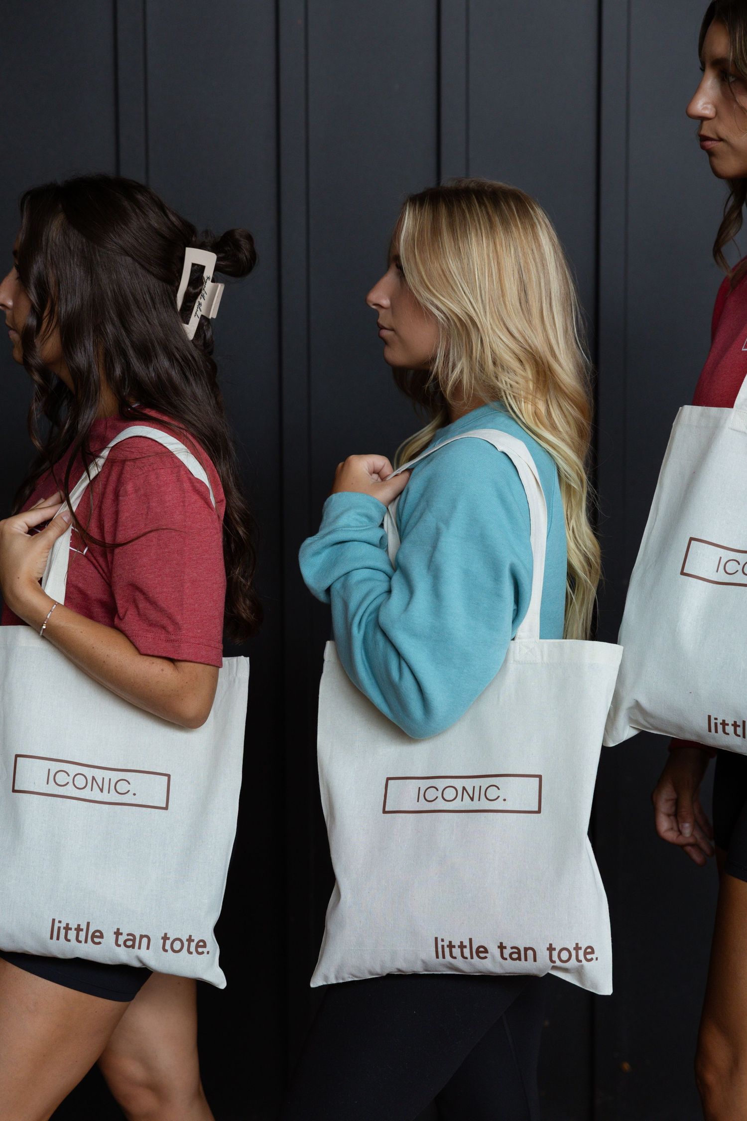 Group holding white tote bags with 'ICONIC' text against dark wall background.