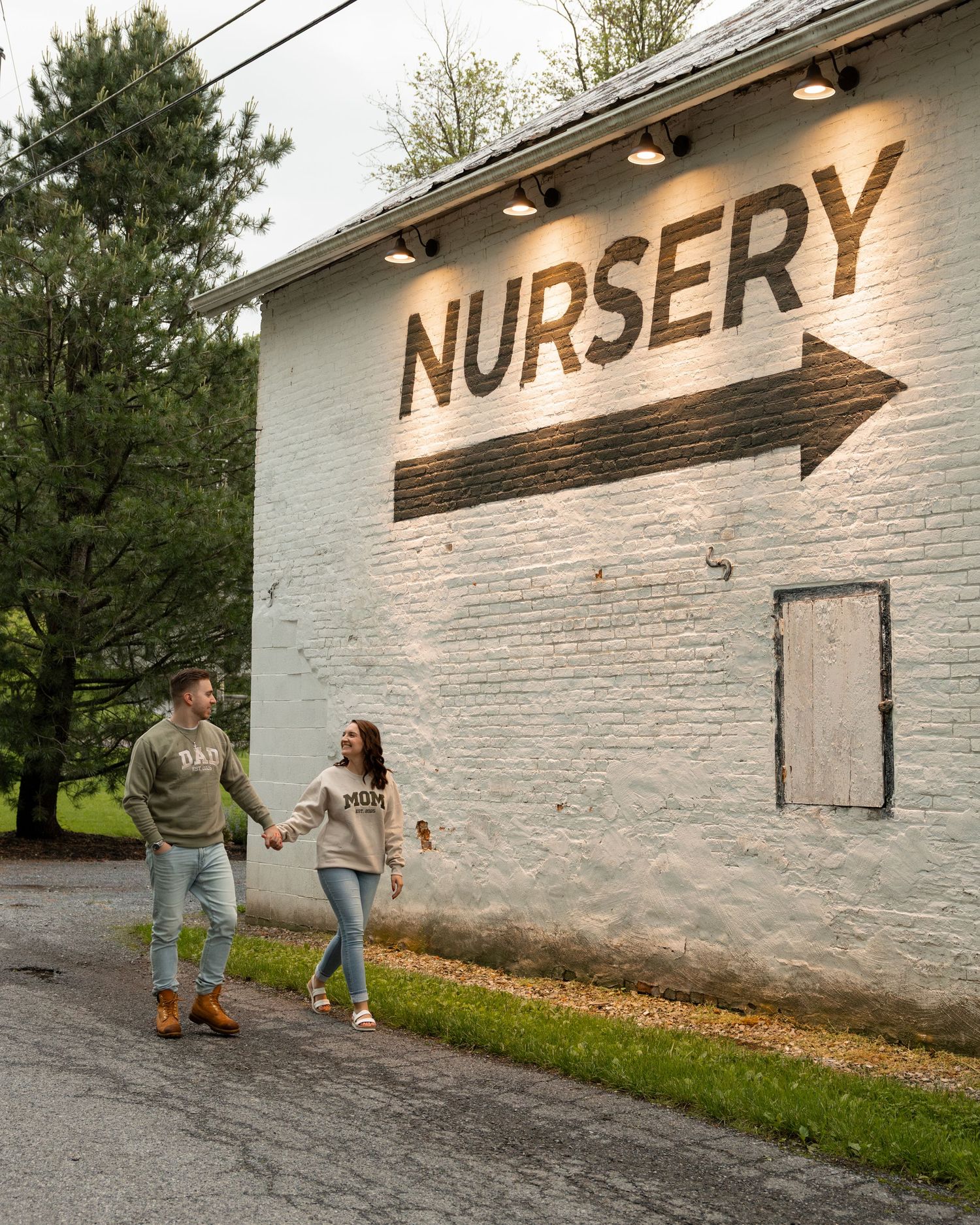 A couple holds hands walking past a white brick building with a large NURSERY sign and arrow painted on the wall.