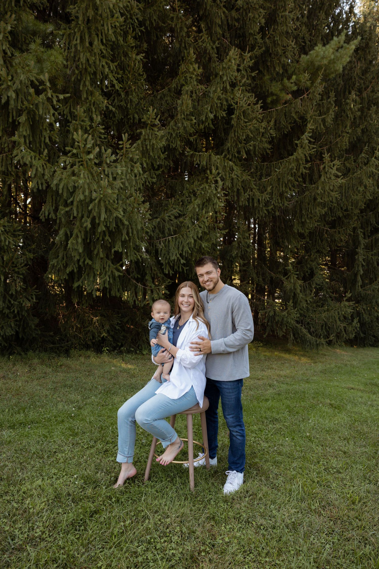 A young family poses outdoors near evergreen trees, with a woman sitting on a stool holding a baby and a man standing beside her.