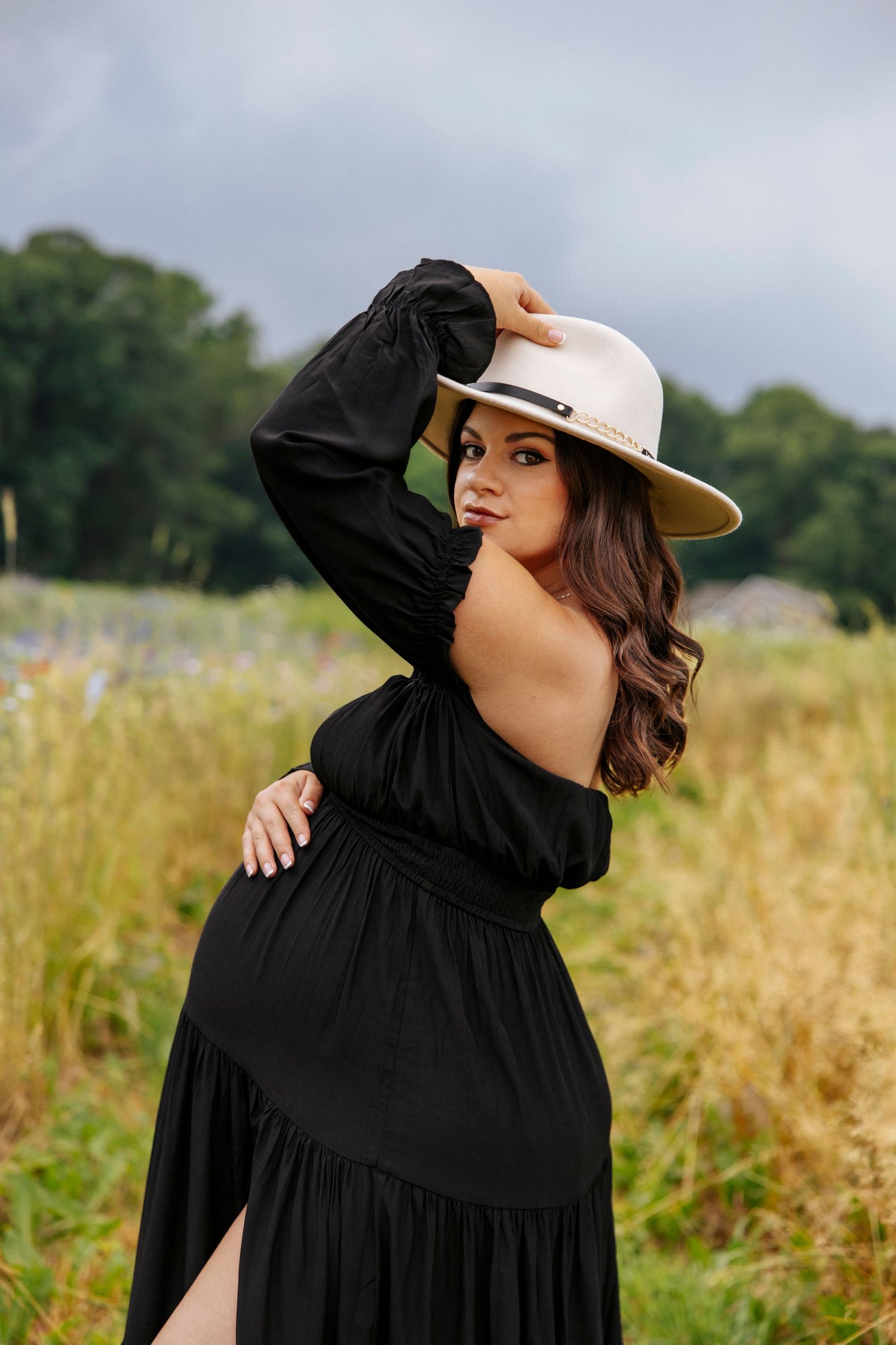 Stylish outdoor portrait in black dress and cream hat against natural field backdrop.