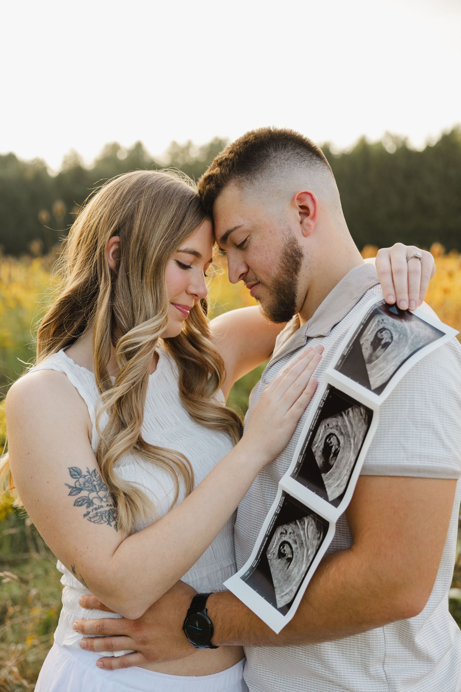 A romantic pregnancy announcement photoshoot featuring ultrasound pictures in a field at sunset.