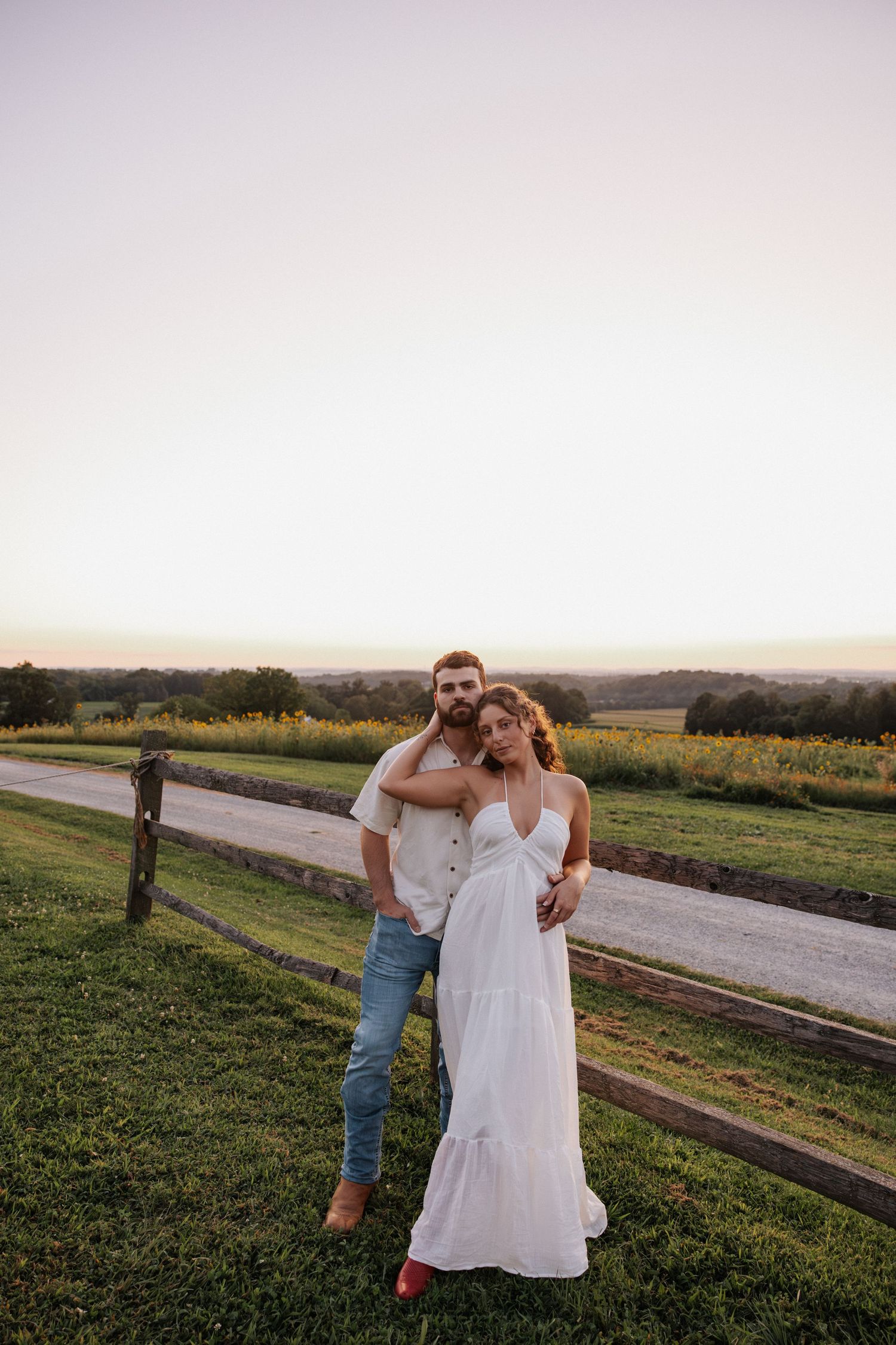 Couple embracing at sunset by a wooden fence on a green farm, woman in white dress, man in jeans and white shirt.