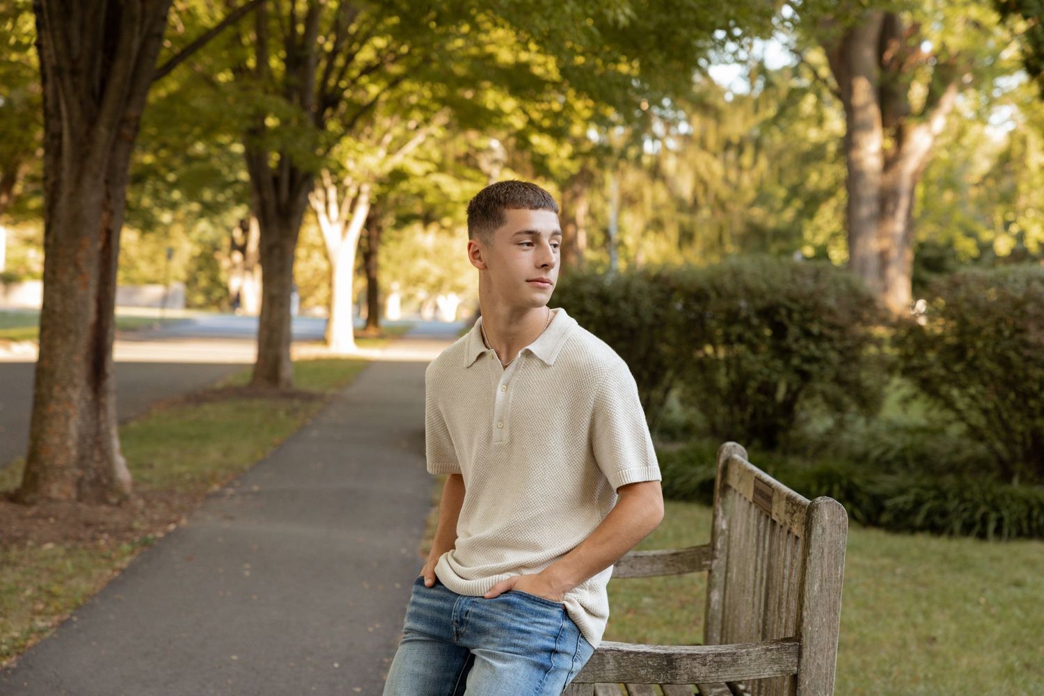 Young man in beige polo and jeans leaning on wooden bench in lush green park along tree-lined path.