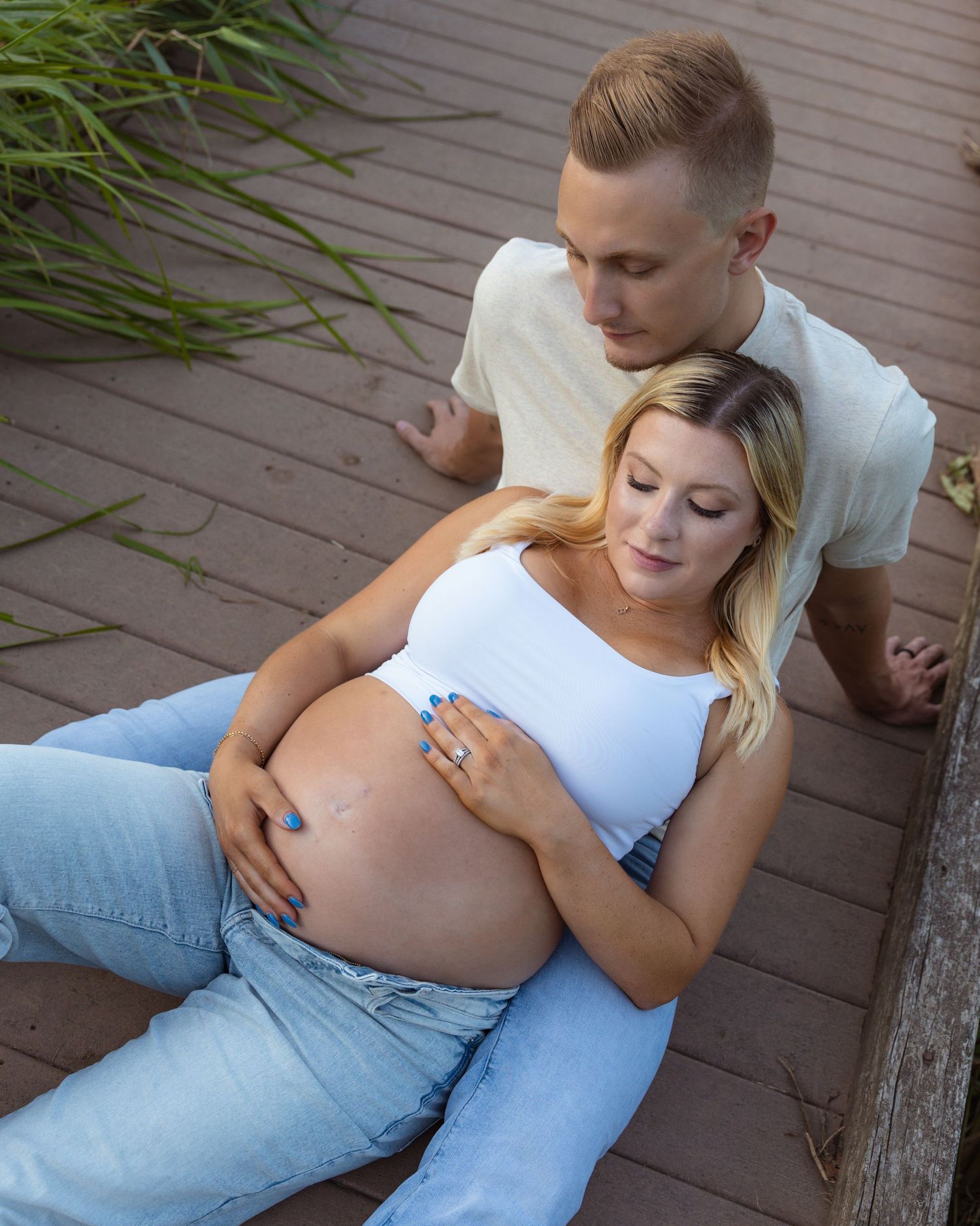Expectant couple in white top and jeans lounging on outdoor deck during pregnancy photoshoot.