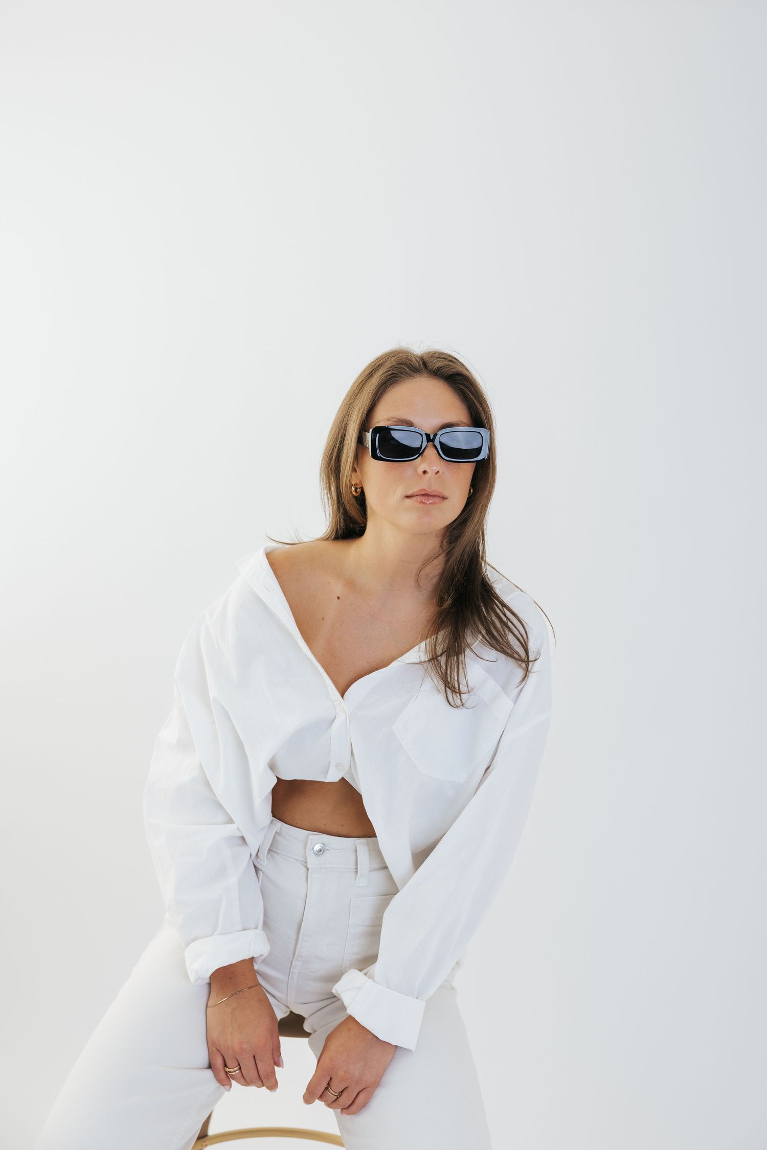 Fashion model in white outfit and sunglasses poses against light background in editorial style photoshoot.