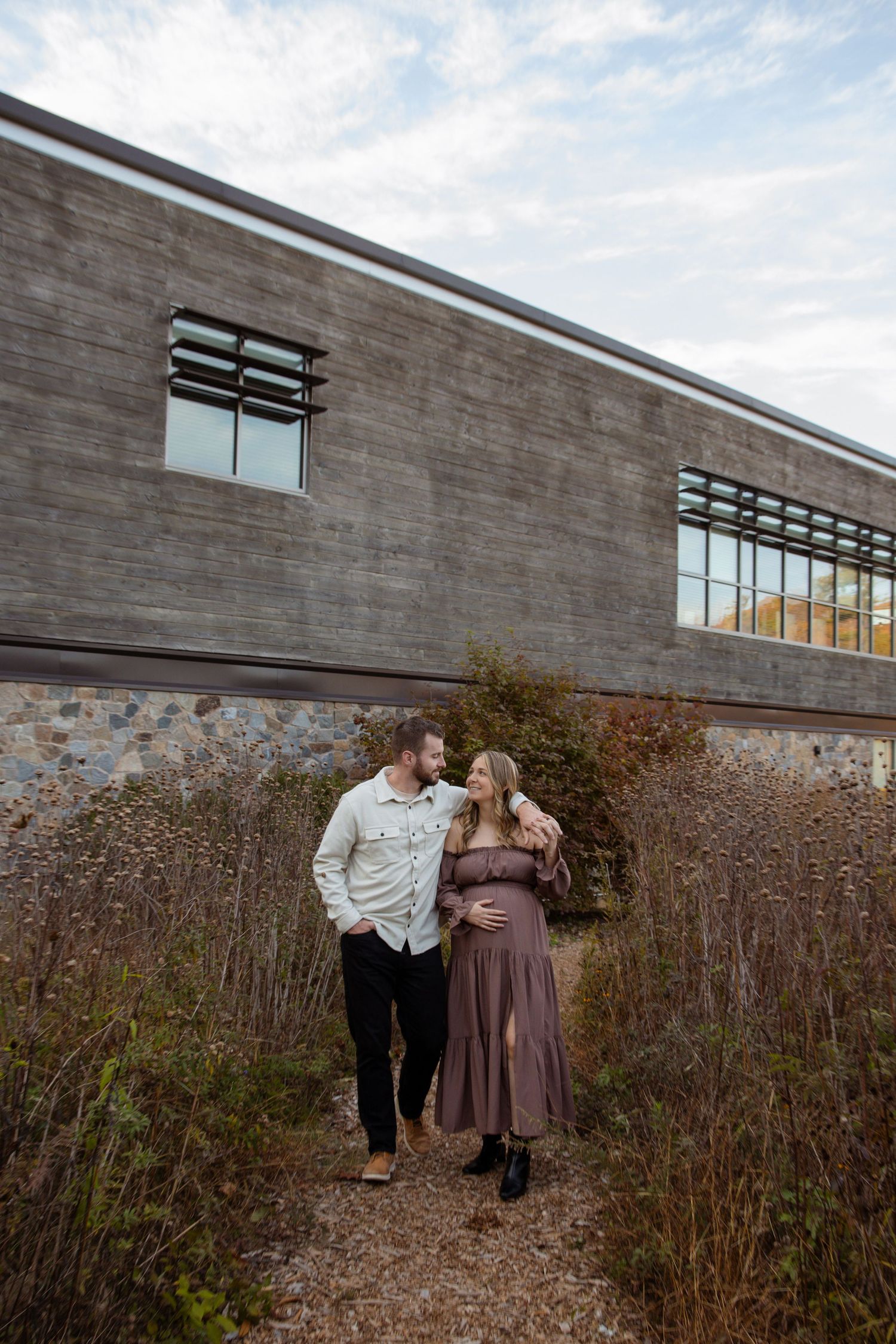 Pregnant couple embracing outdoors near modern stone building surrounded by tall wild grasses under a cloudy sky.