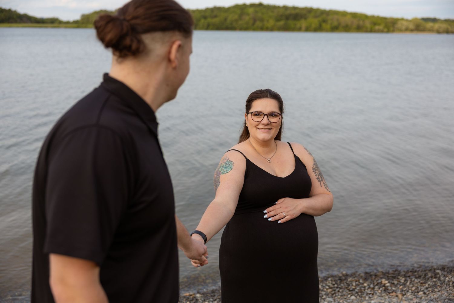 A couple shares tender moments during a lakeside photoshoot with water and trees in view.