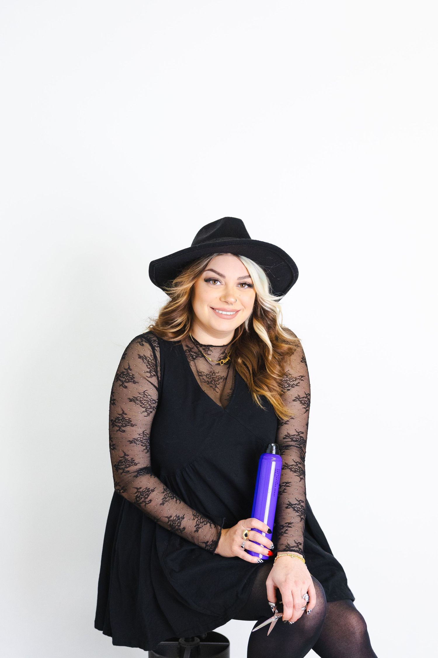 Model in black dress and hat holding purple can against white backdrop.