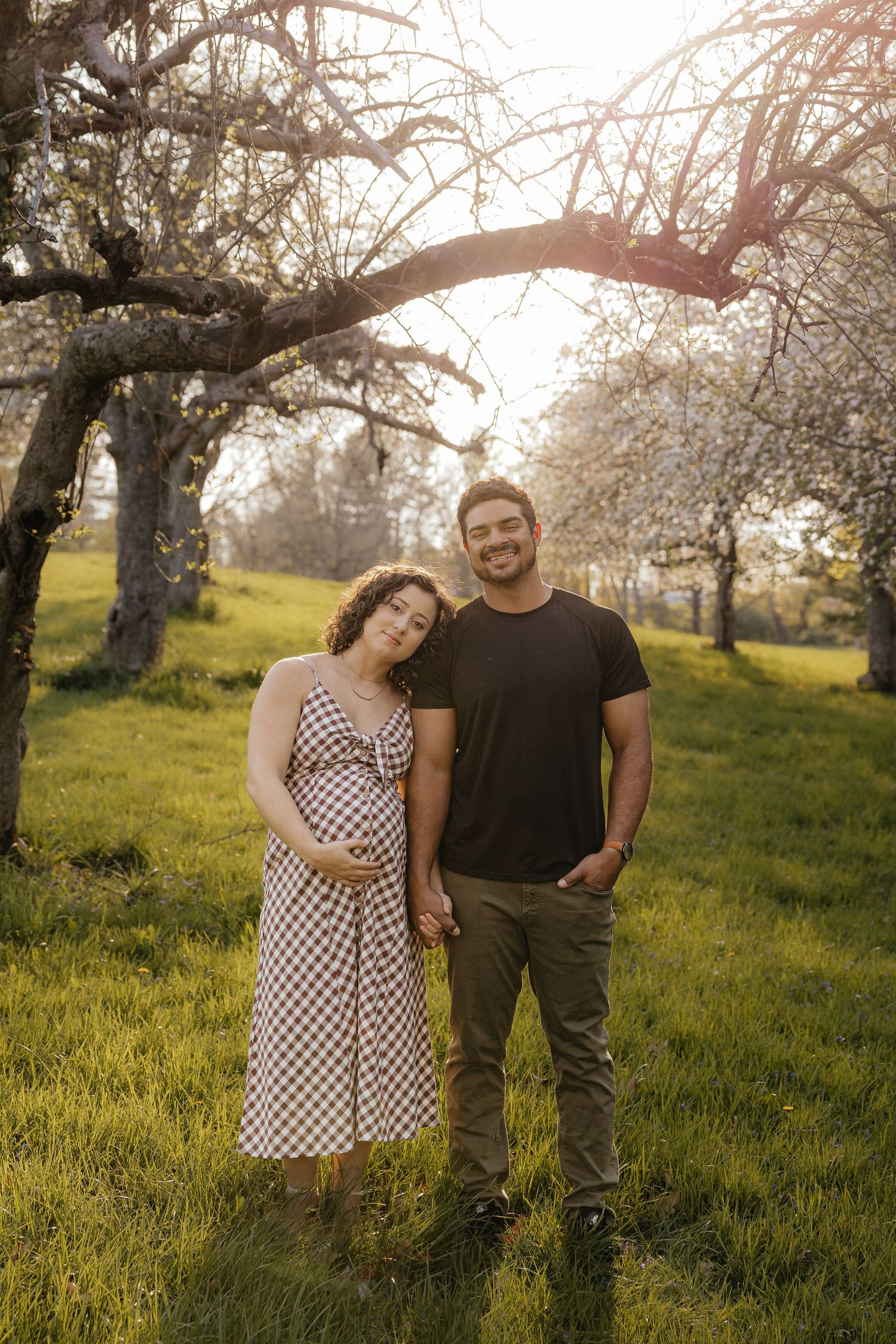 Pregnant couple holding hands in sunlit orchard with blossoming trees and green grass during golden hour.