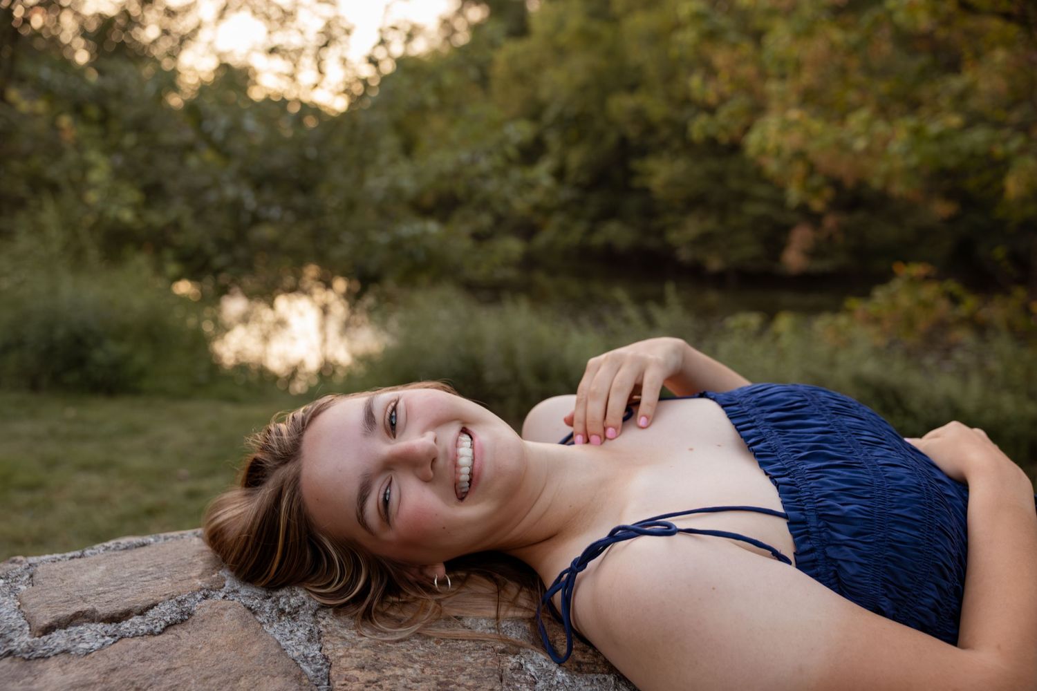 A smiling woman in a navy blue bikini top lying on a rock outdoors, surrounded by lush green trees.