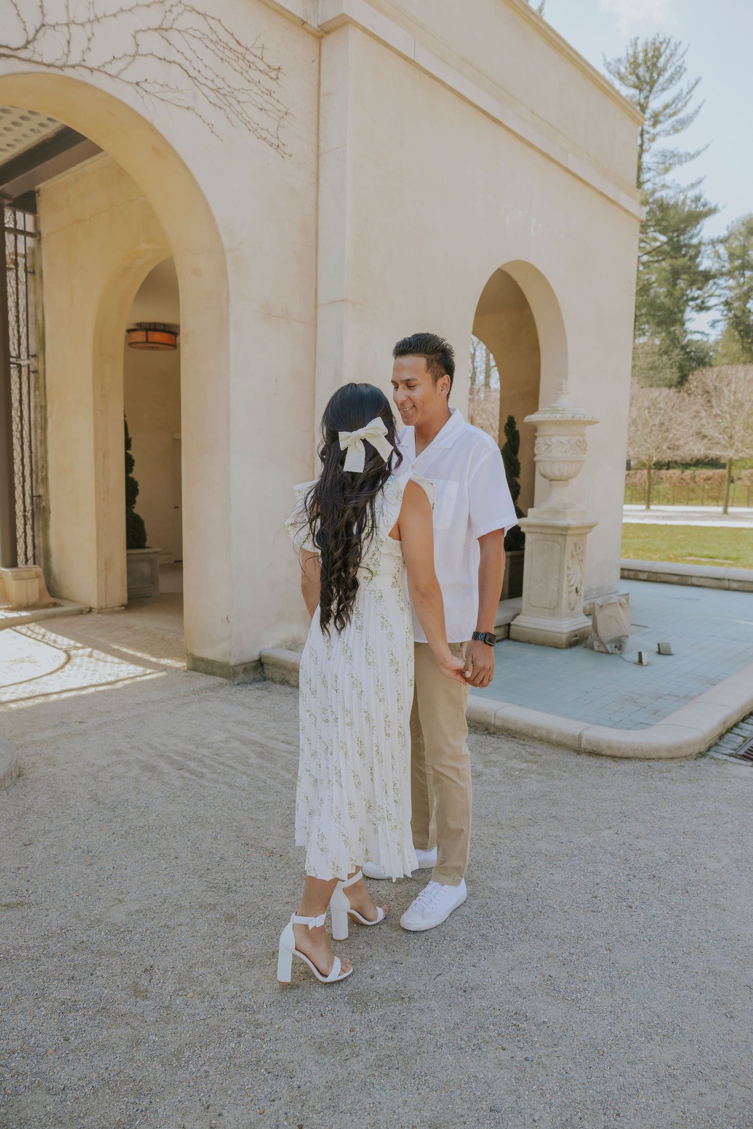 Couple in coordinated white and beige outfits pose along stone colonnade walkway.