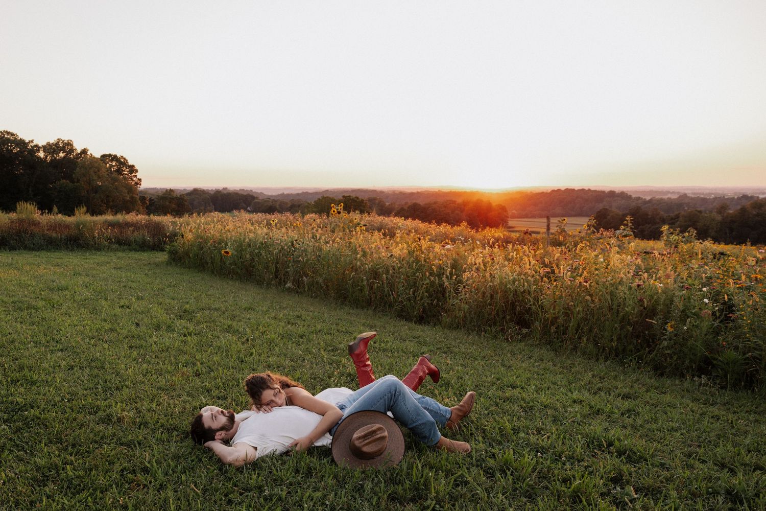 Couple lying on green grass at sunset near a wildflower field in a romantic countryside setting.