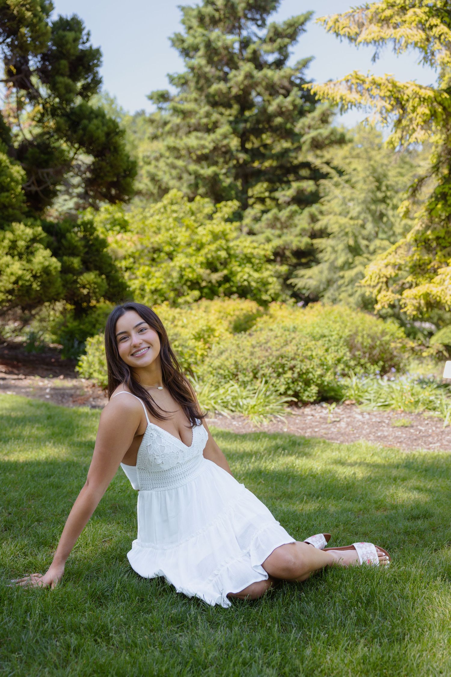 Figure in white dress reclines on grass in a sunlit garden with evergreen trees.