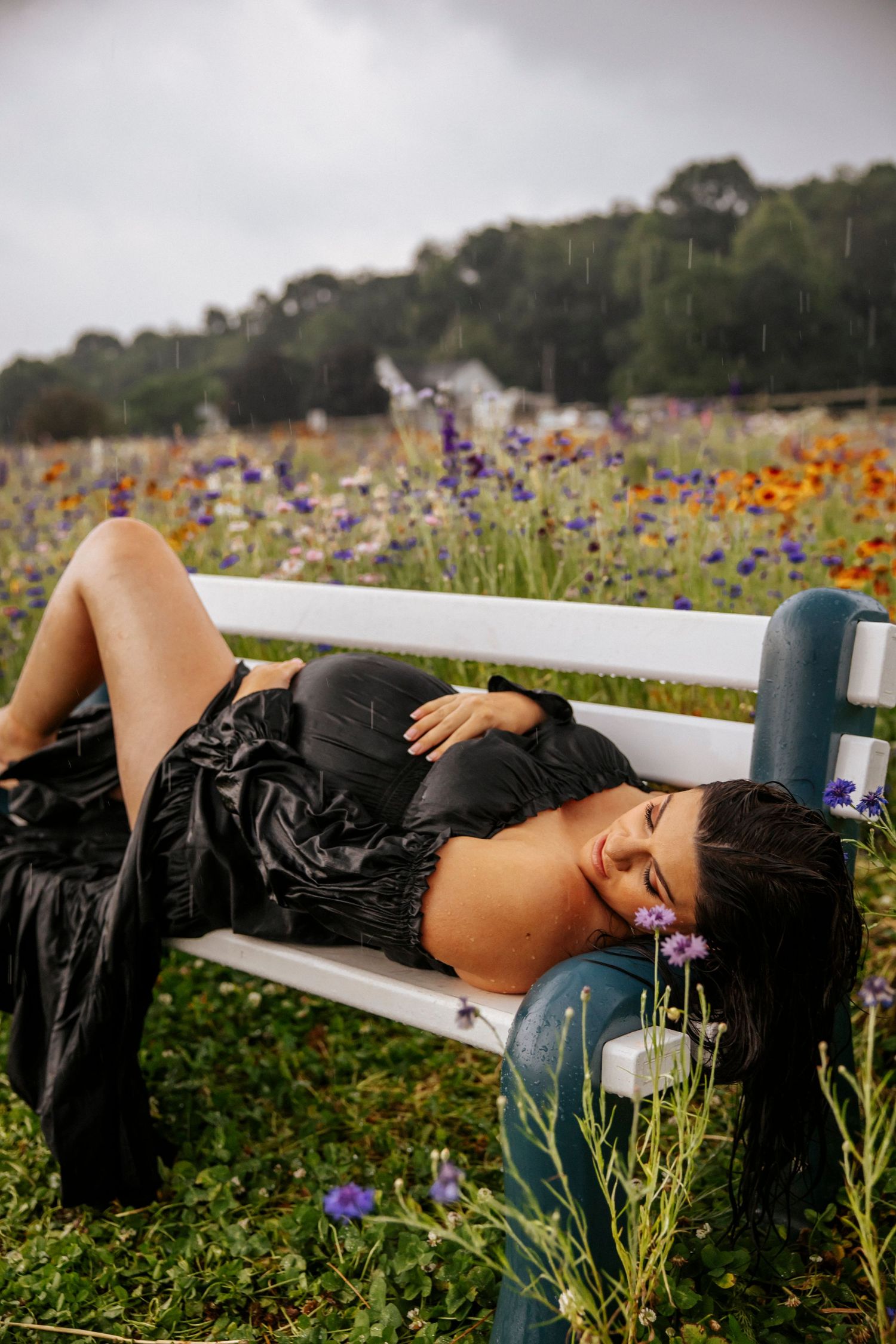Figure in black dress reclines on white fence amid blooming wildflower field.