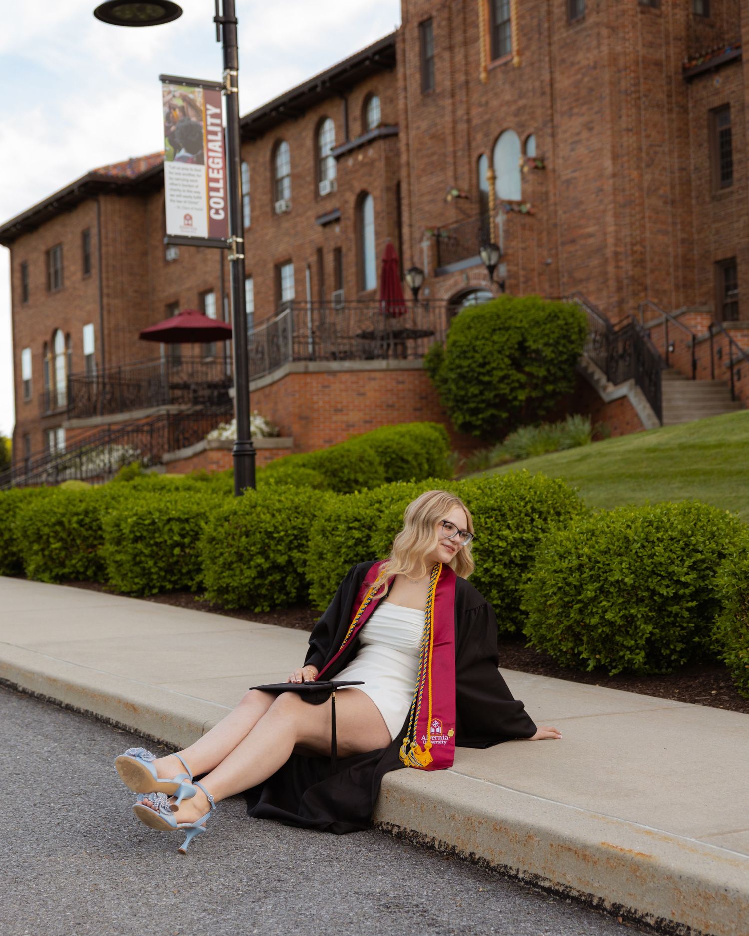 Graduate in academic dress poses casually on campus grounds near brick university building.
