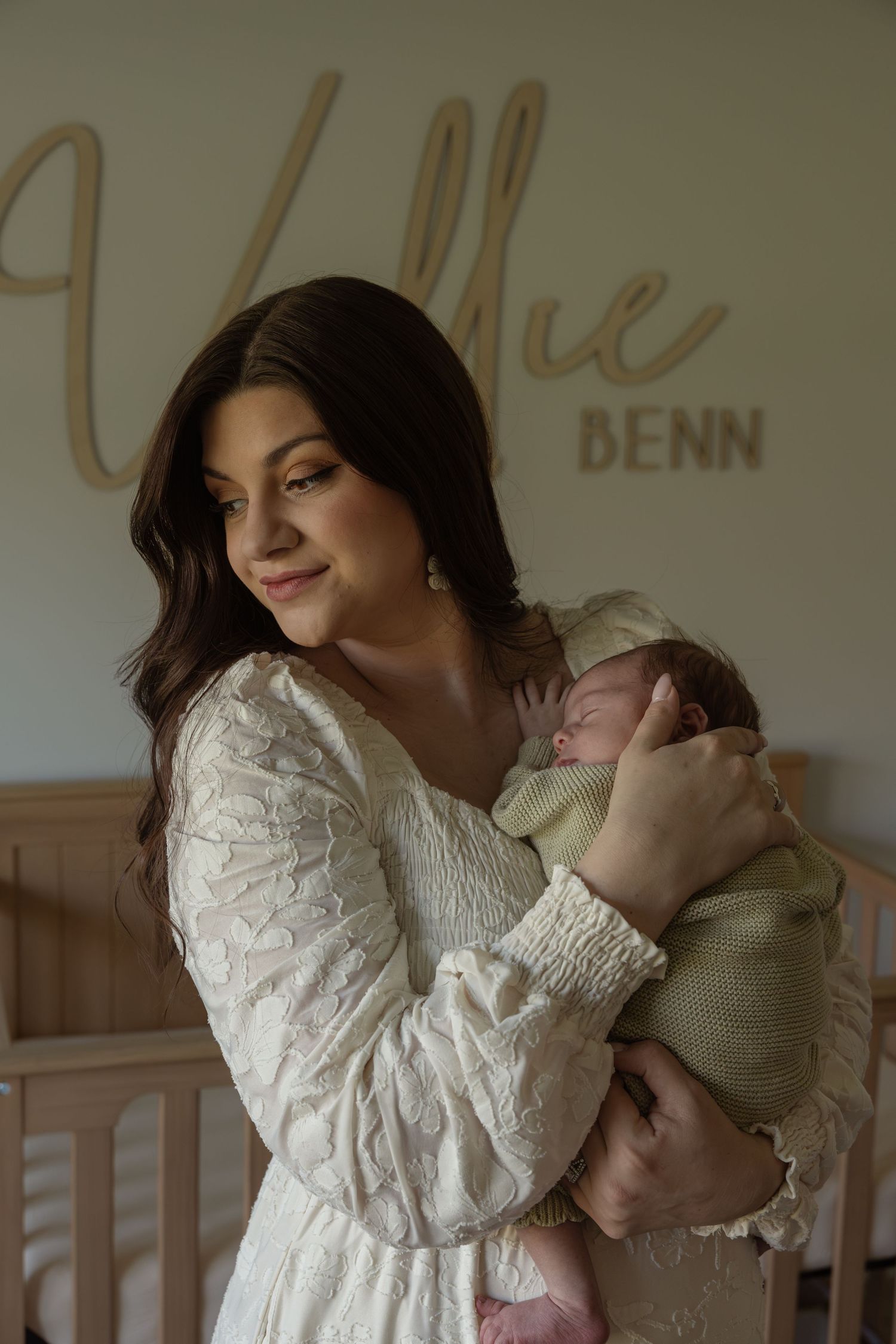 A mother in a white lace dress tenderly holds her newborn baby in a nursery with a name sign reading Vallie Benn.