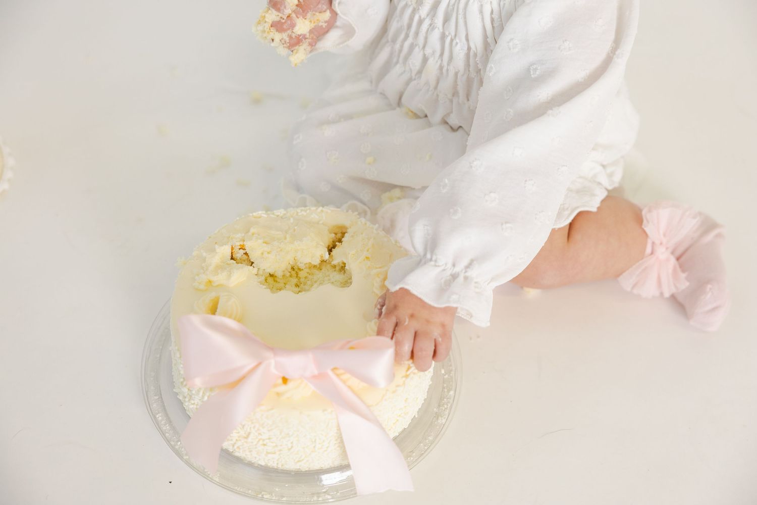 Aerial view of white cake with pink bow and small hand reaching for it.