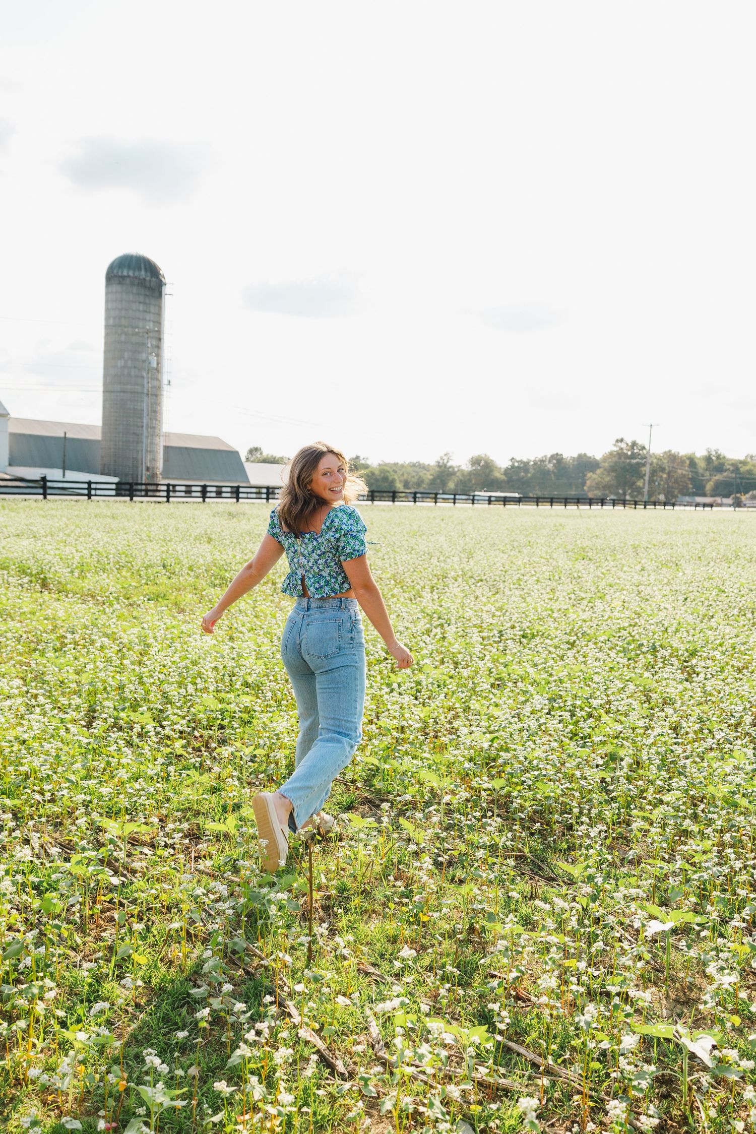 Person in blue outfit twirls through field of white wildflowers near rural barn.