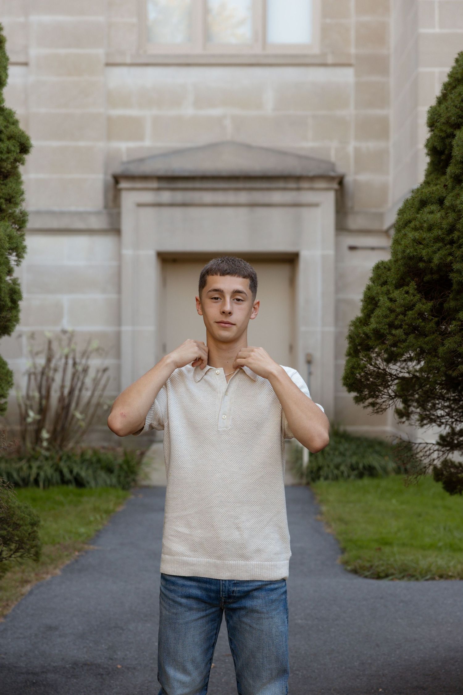 Young man in beige polo shirt and jeans posing confidently outdoors in front of a stone building with green shrubs.