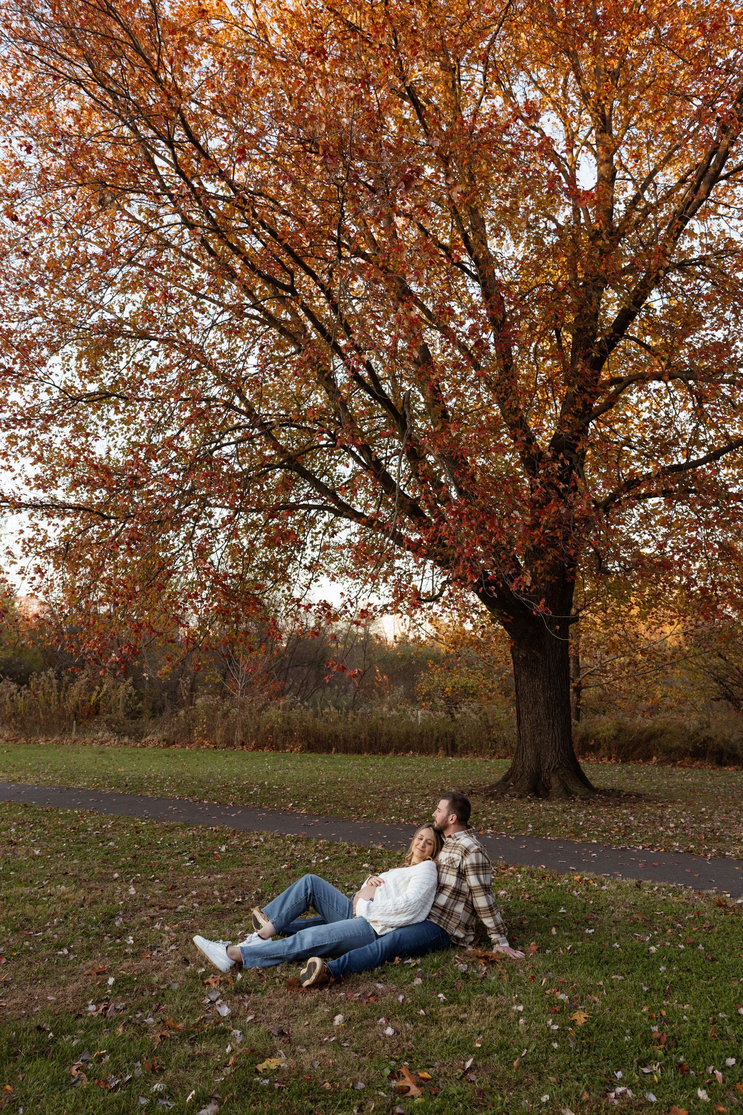 Couple relaxing under a vibrant autumn tree in a park, surrounded by fallen leaves on a peaceful fall day.