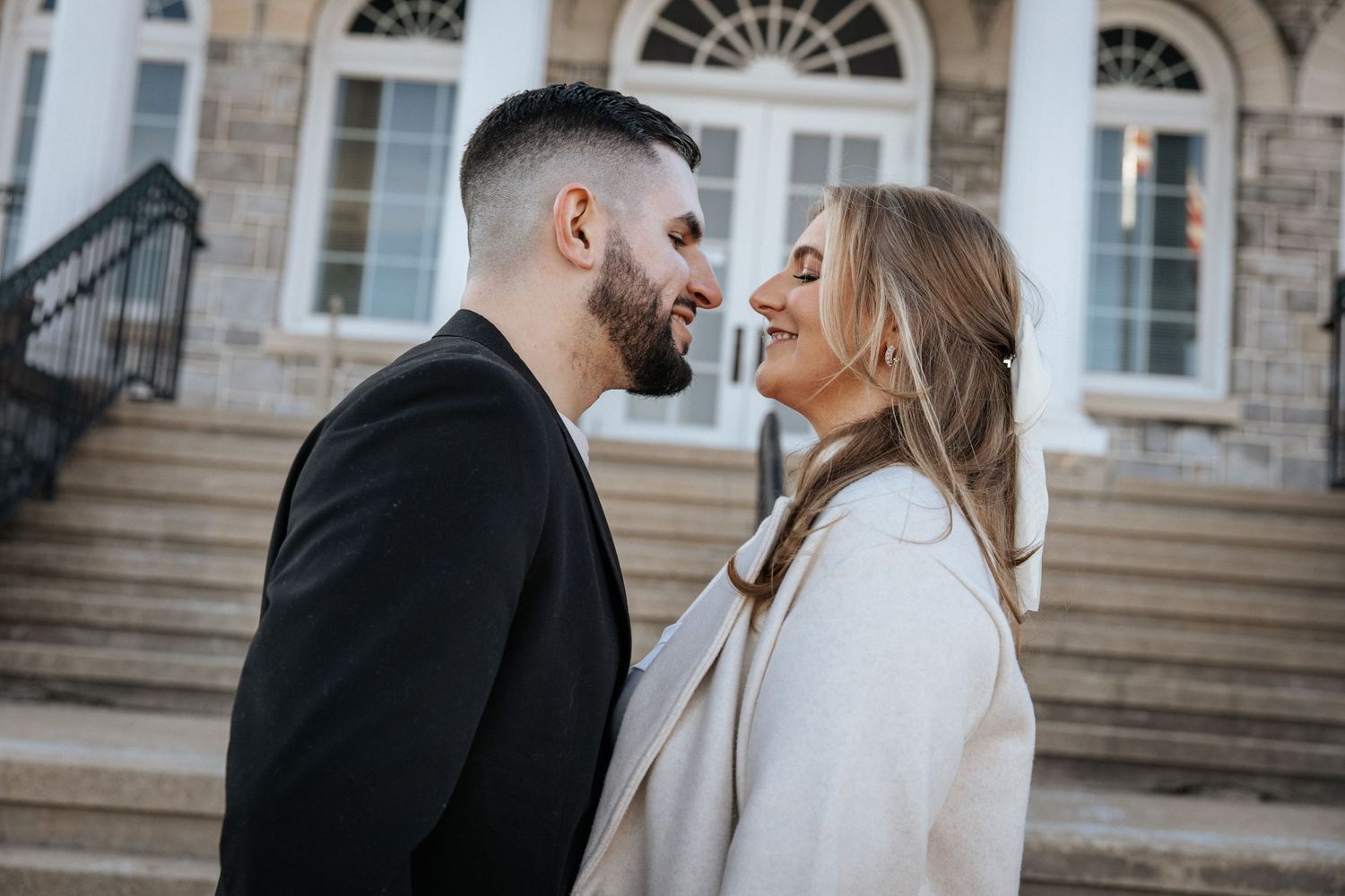 A romantic couple embraces on the steps of a stone building with elegant architectural details.