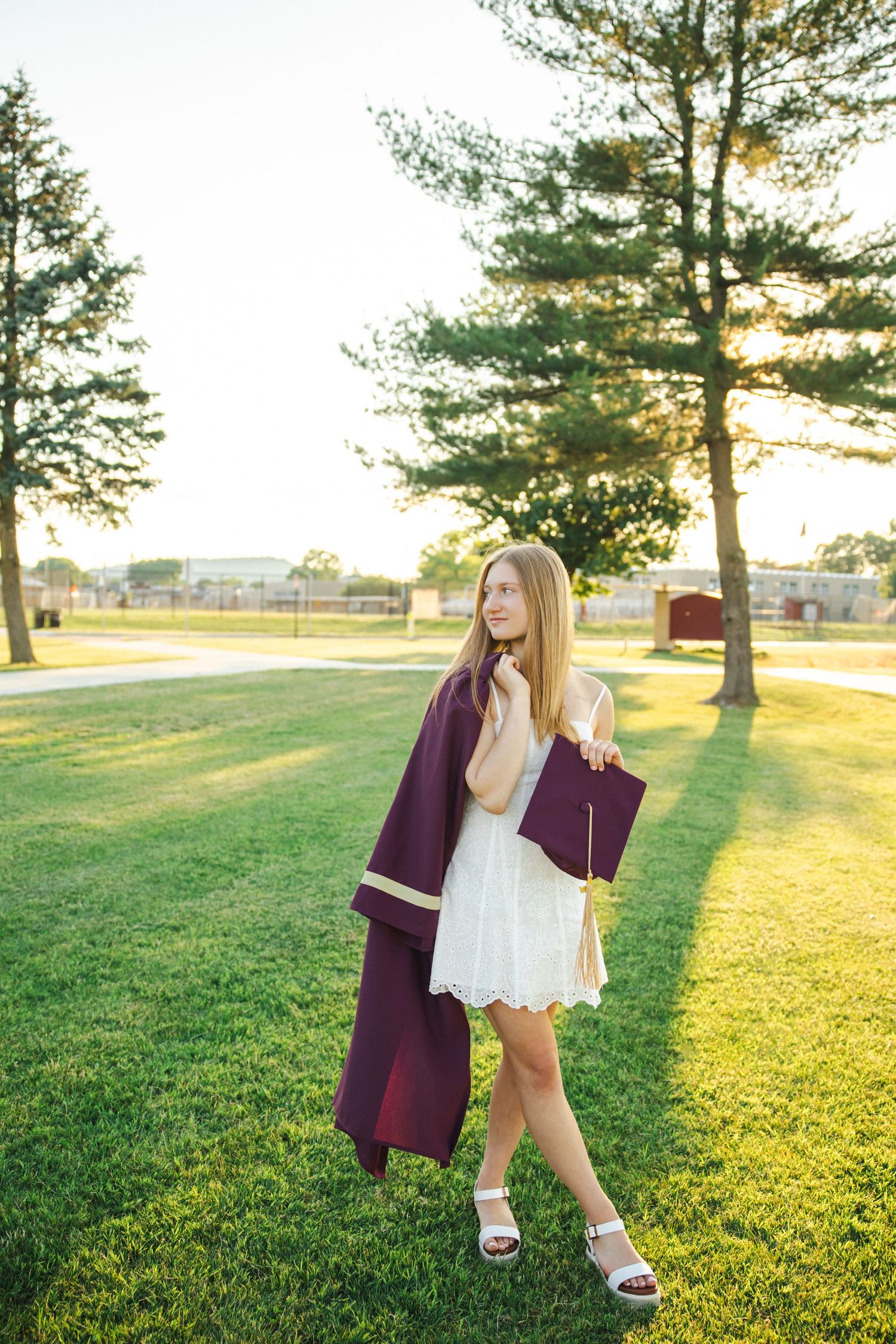 Graduate in white dress carries maroon graduation gown while walking through sunlit campus.