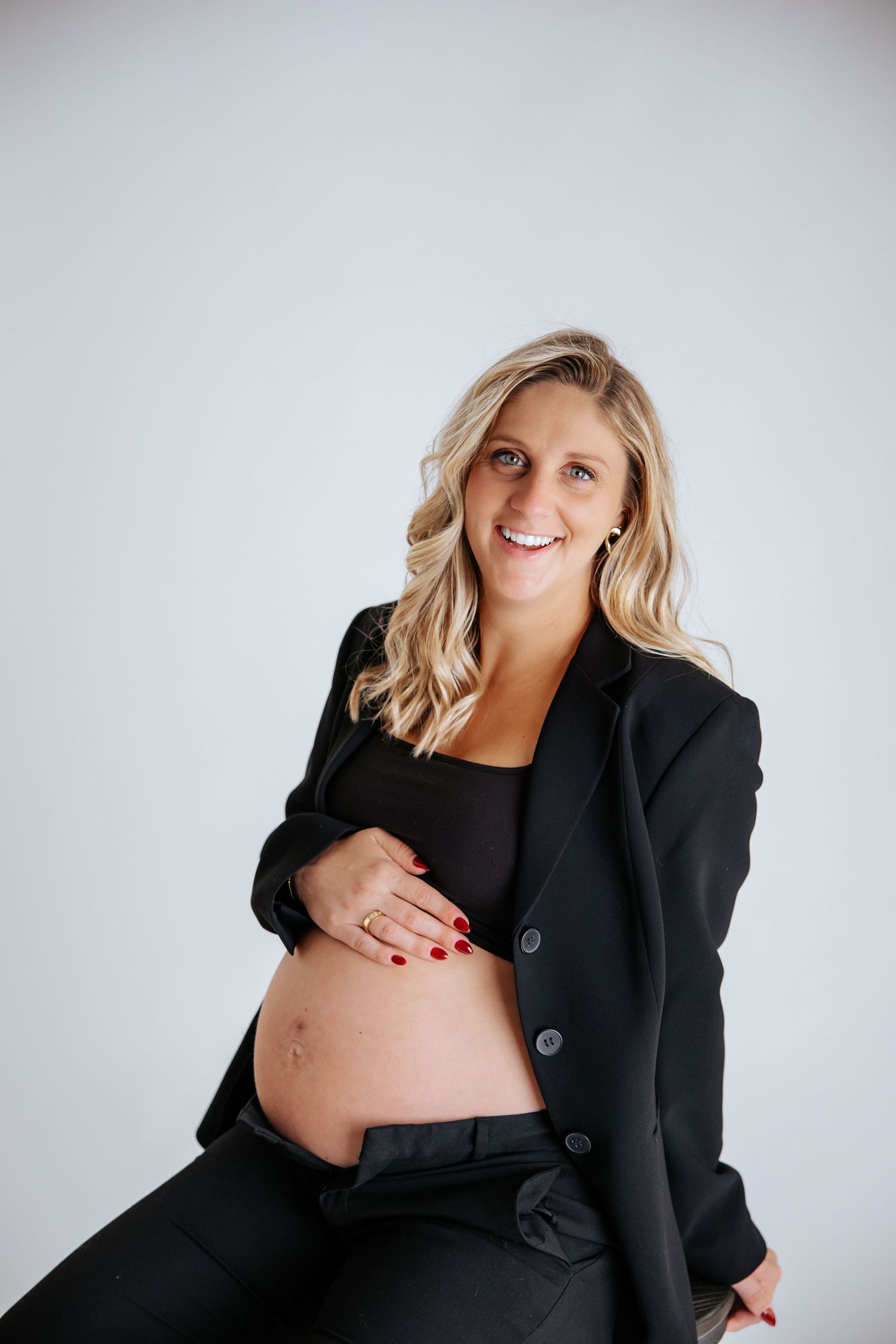 Professional maternity photo with woman in black blazer seated against white background.