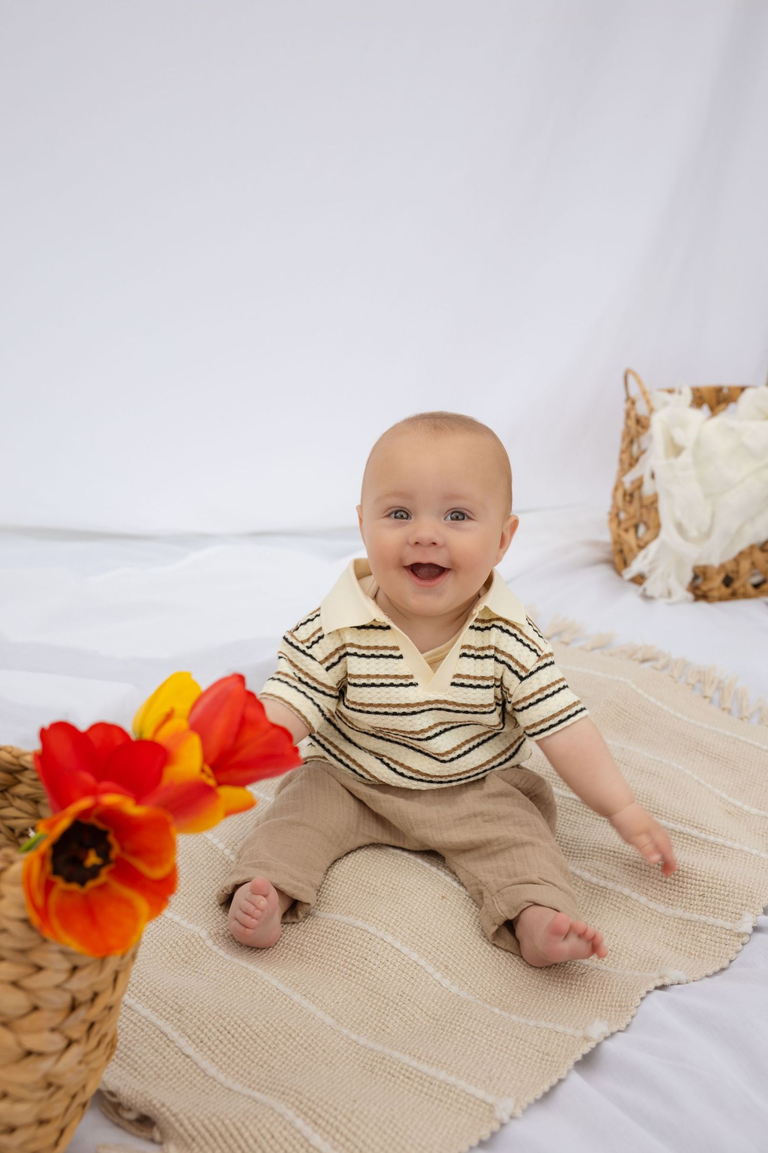 Happy baby in striped shirt posed with bright orange poppies on cream blanket.