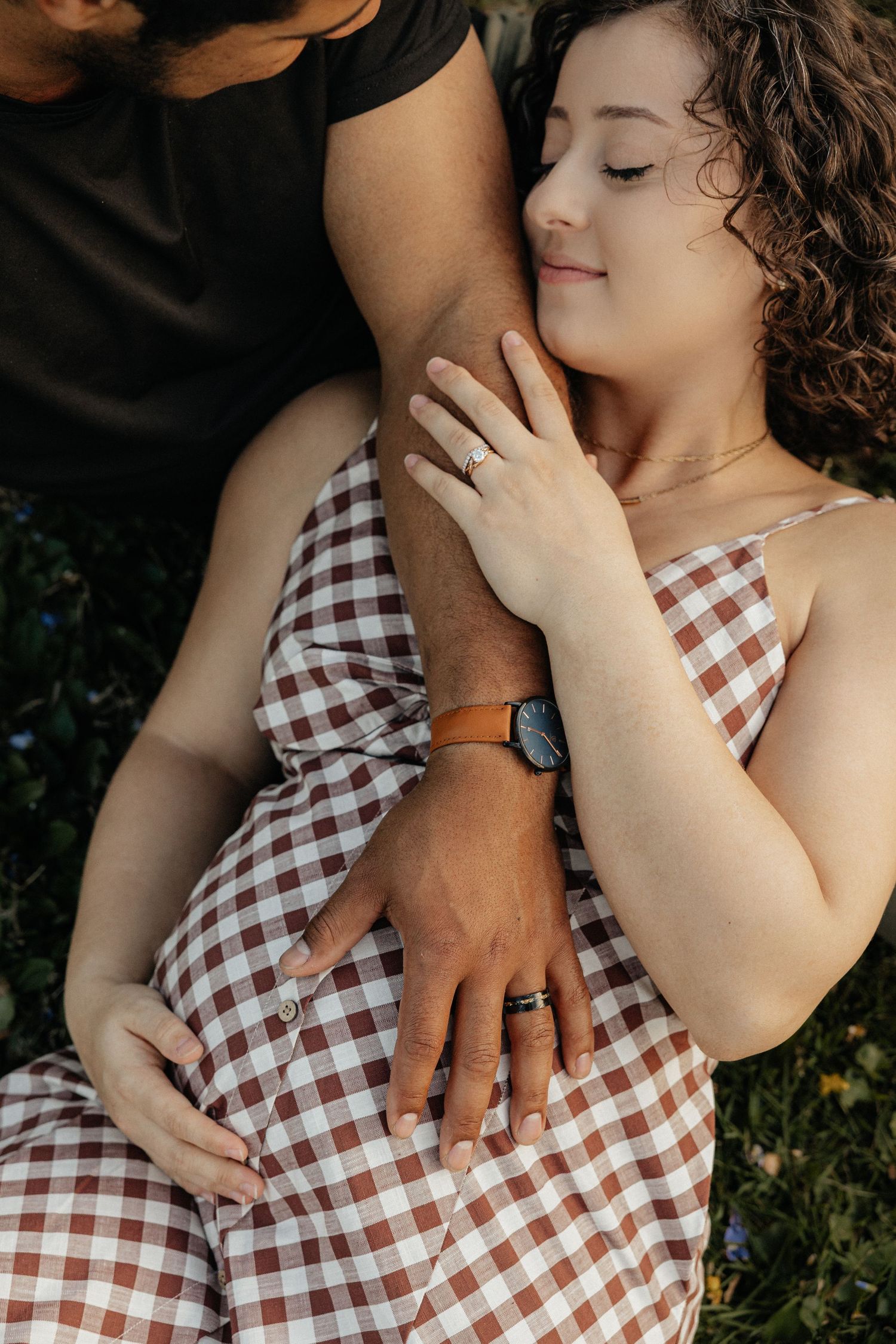 Romantic couple relaxing outdoors, woman in plaid dress with eyes closed, man's arm around her, both wearing rings.