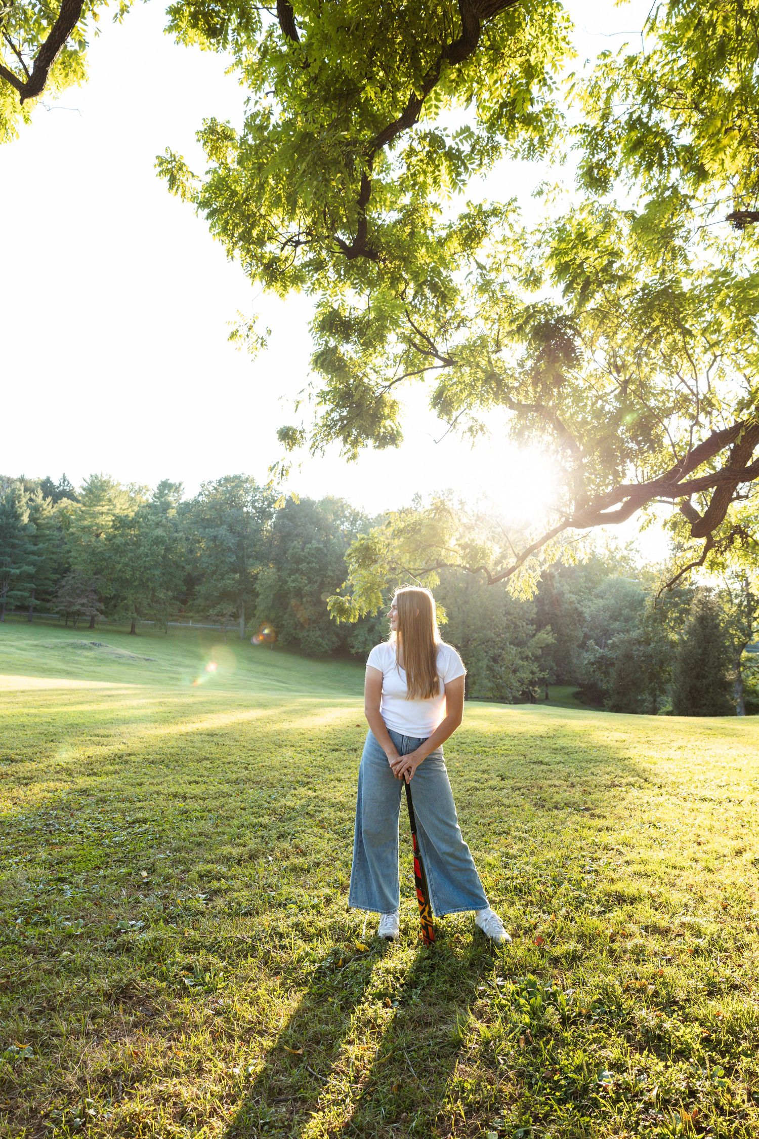 Young woman in white top and jeans stands on lush green grass holding a golf club at sunset in a park.