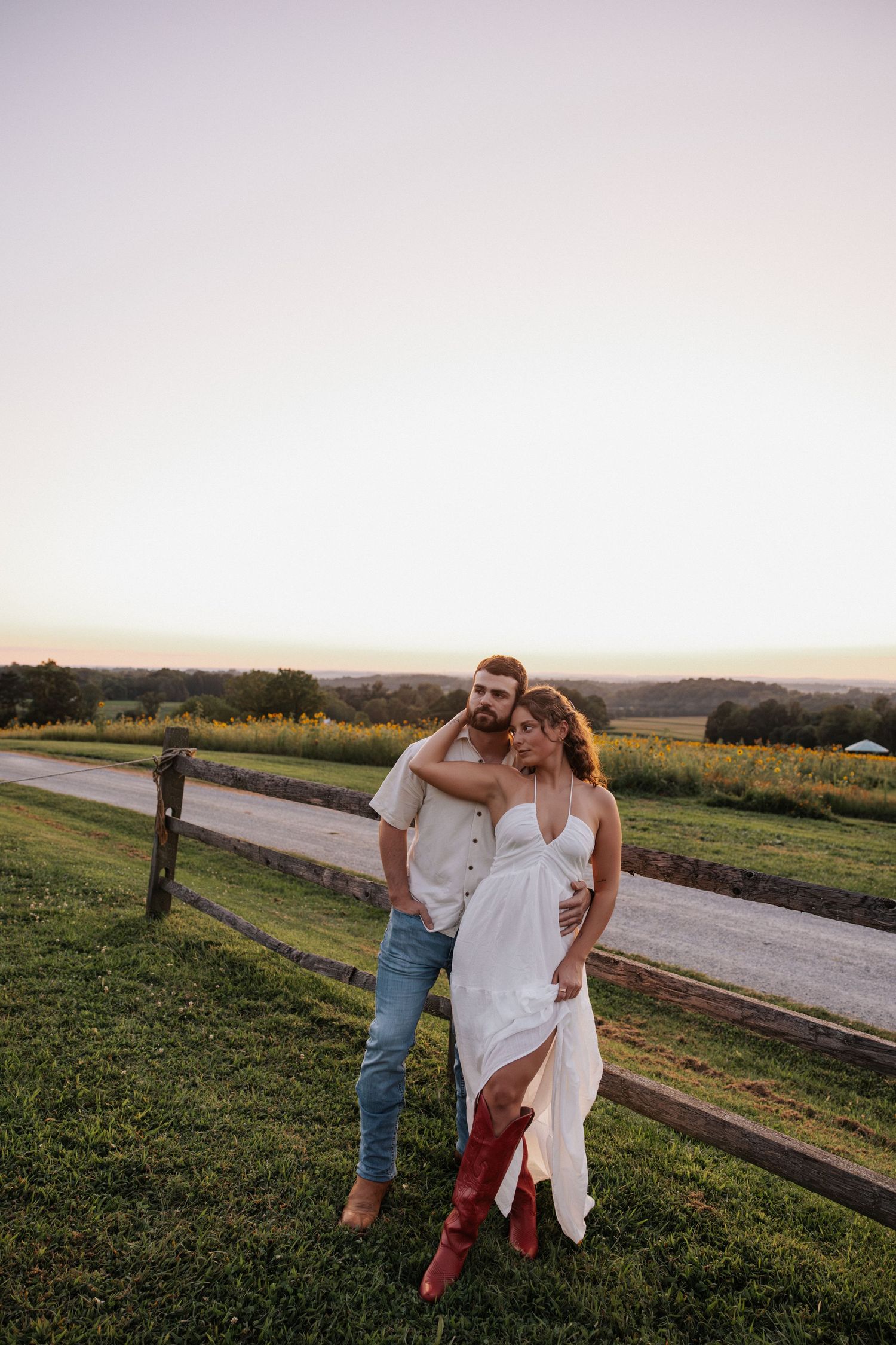 Couple in white outfits embracing on grassy hill at sunset near country road.