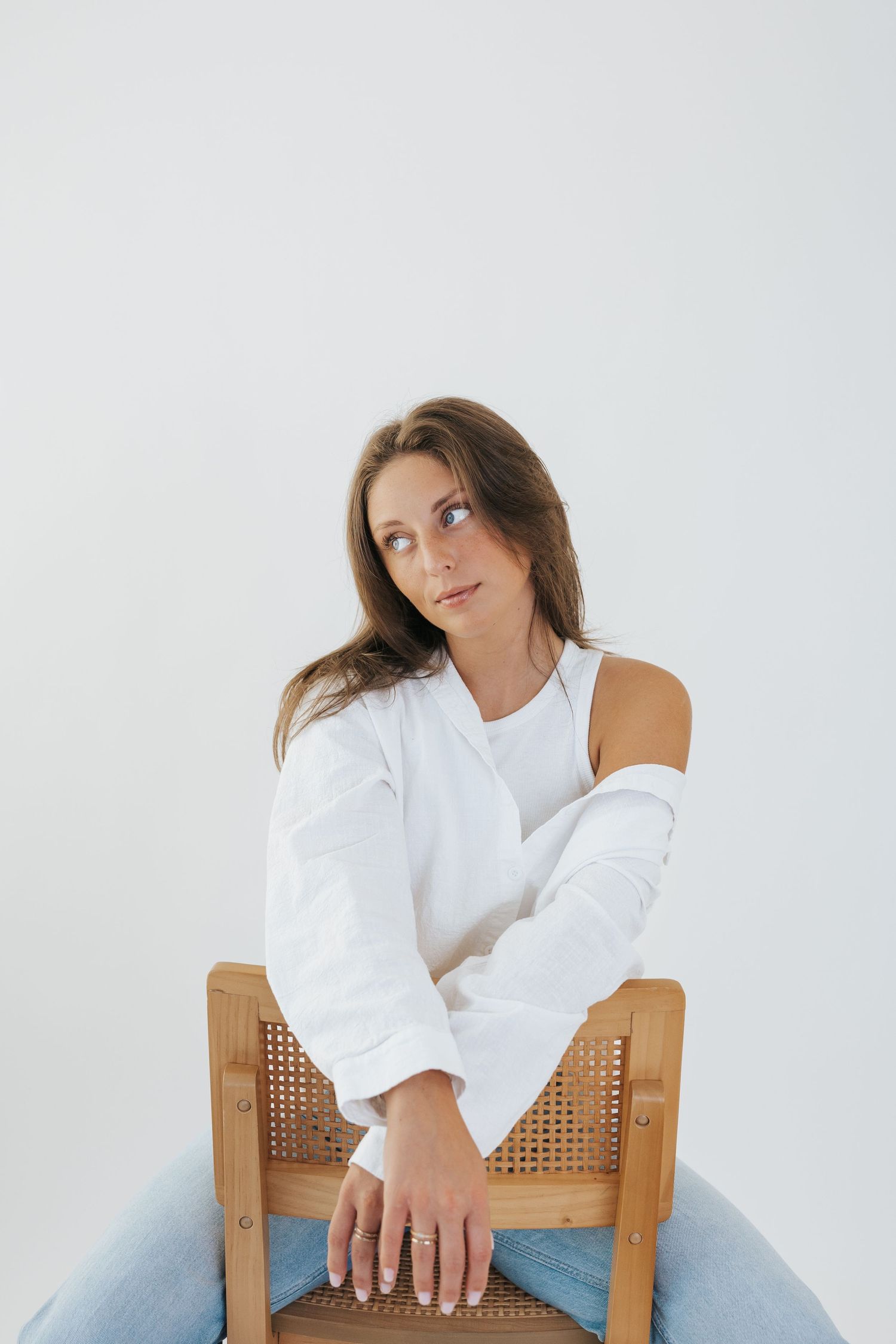 Model wearing white top poses on wooden chair in minimalist studio setting.