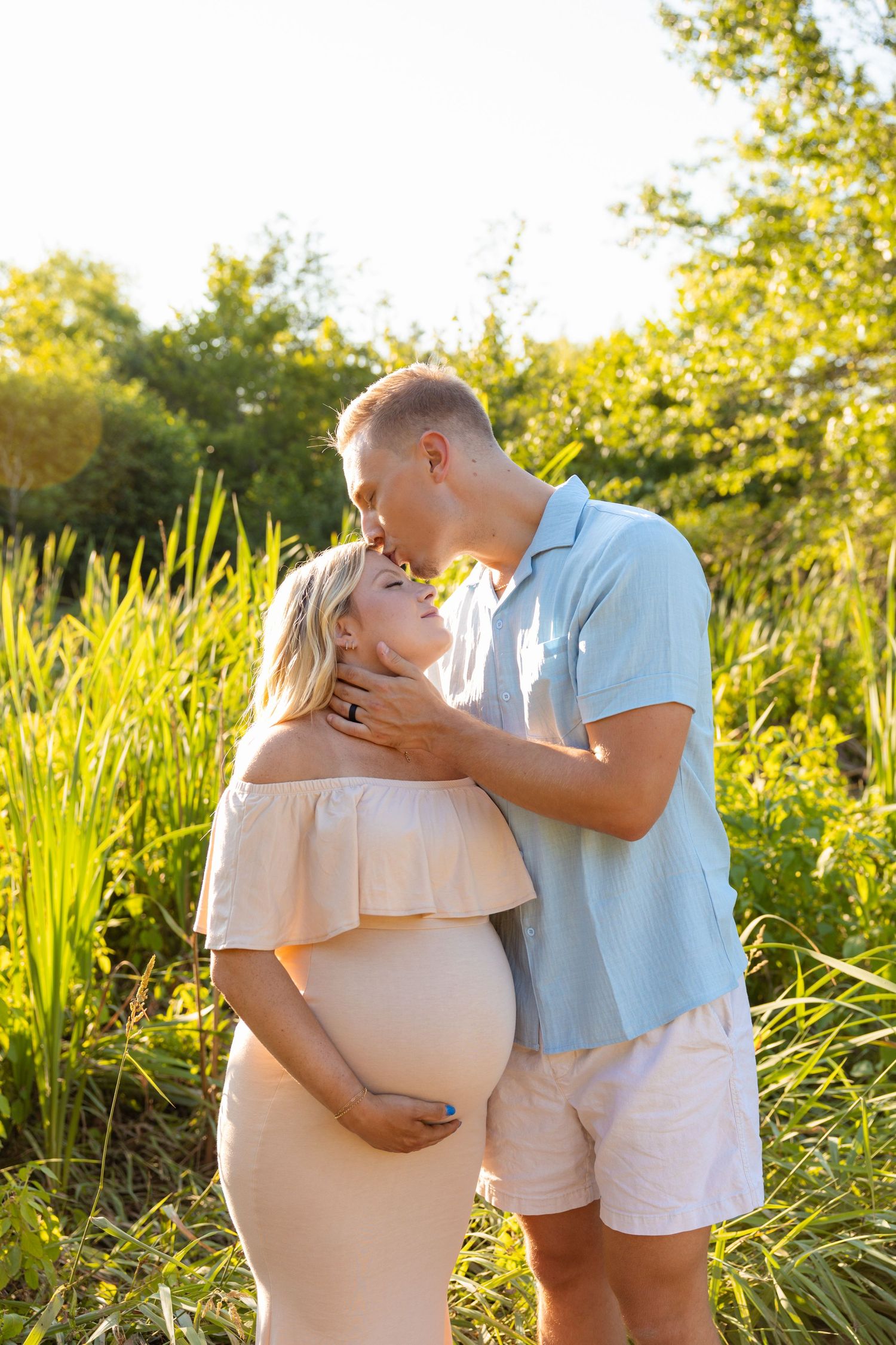 Couple embraces in golden sunlight among tall grasses during maternity photoshoot.