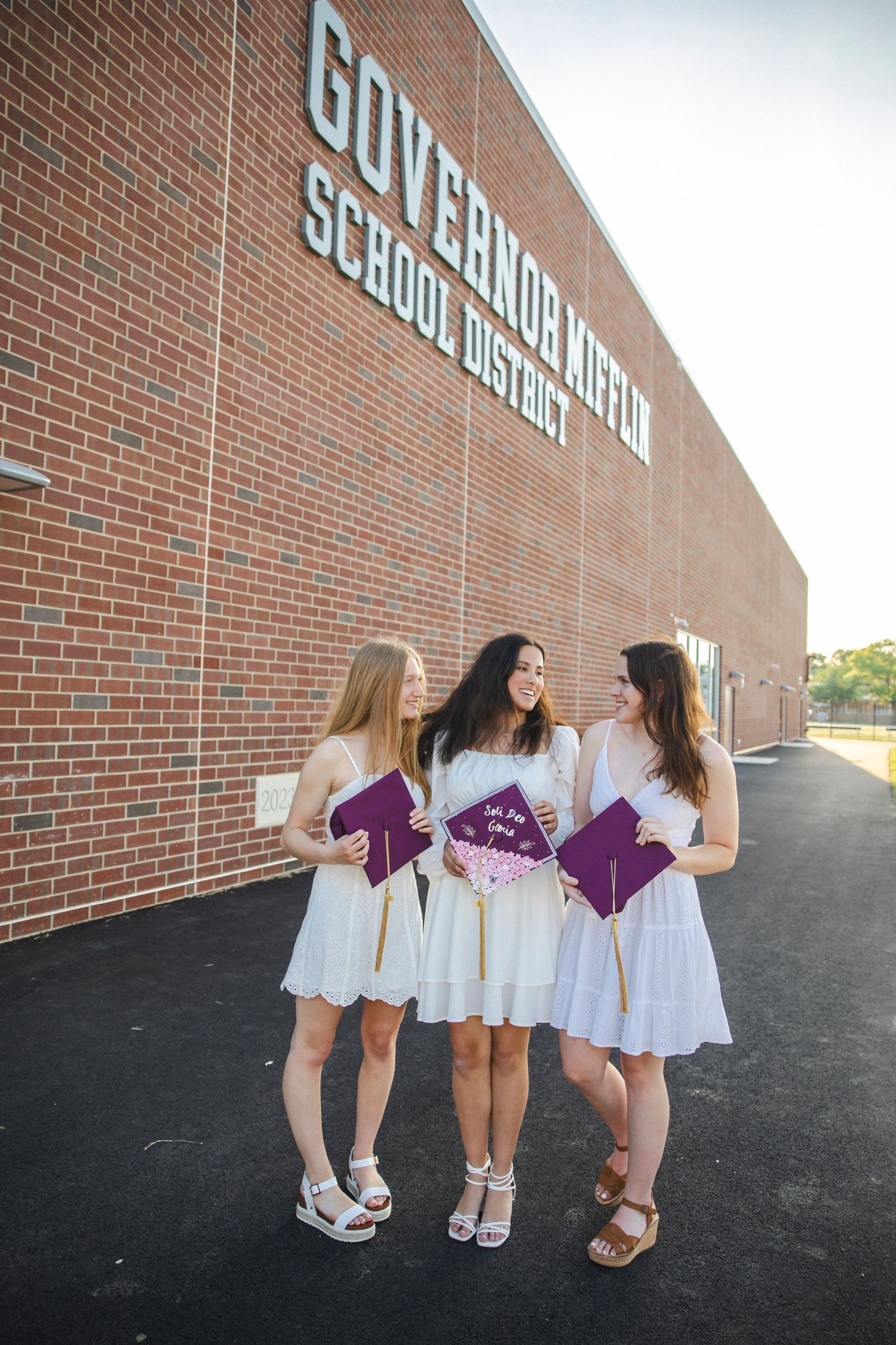 Three graduates in white dresses hold diplomas while posing by brick school building.
