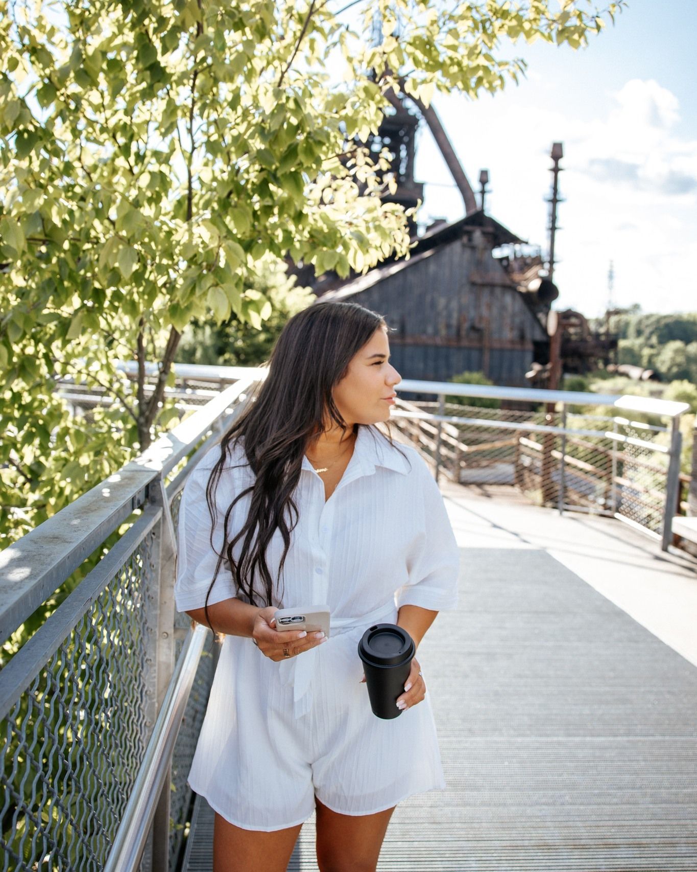Woman in white romper holding coffee cup and phone on a wooden bridge surrounded by green trees.