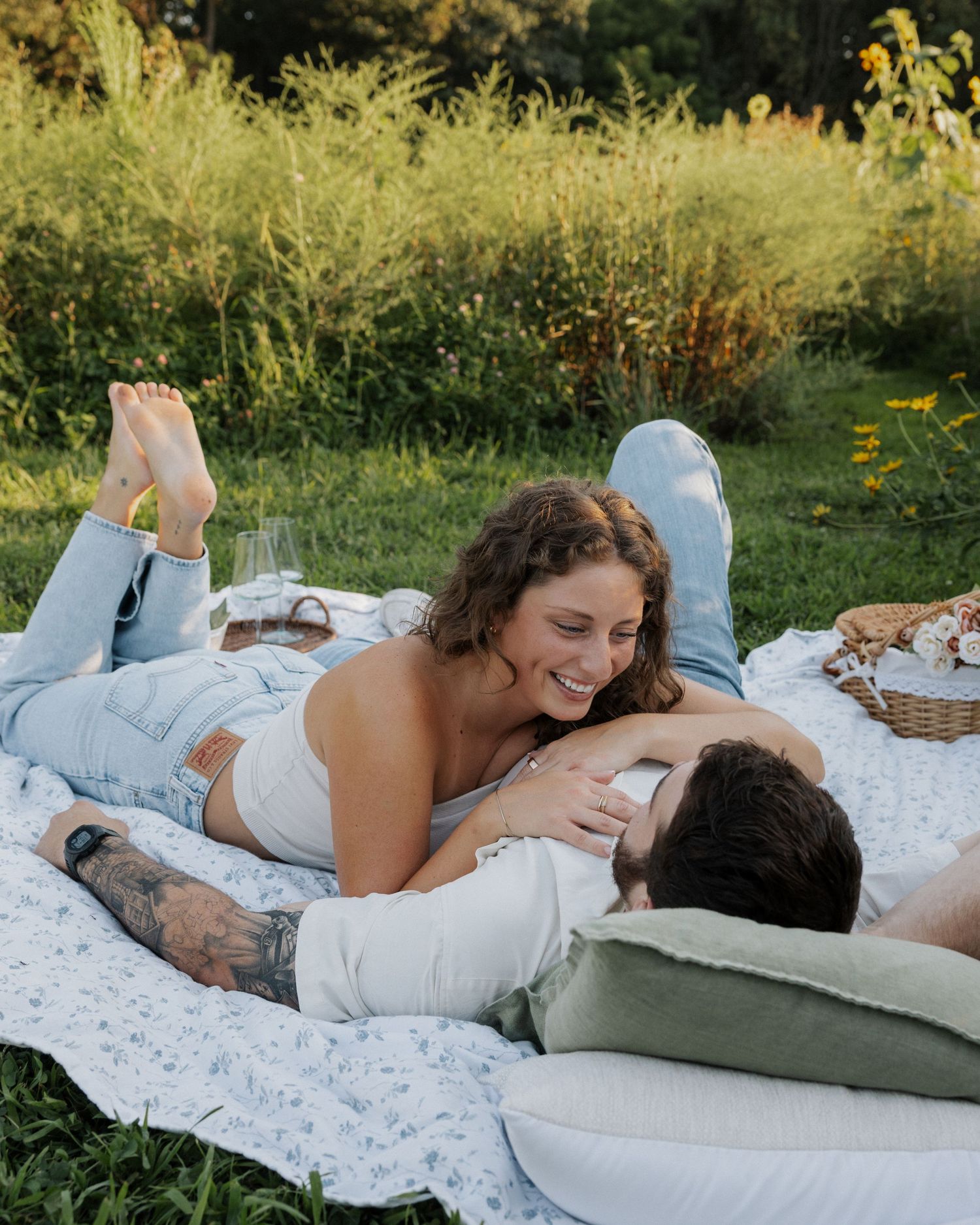 A couple laughs and cuddles on a floral picnic blanket in a lush green meadow, enjoying a romantic outdoor picnic.