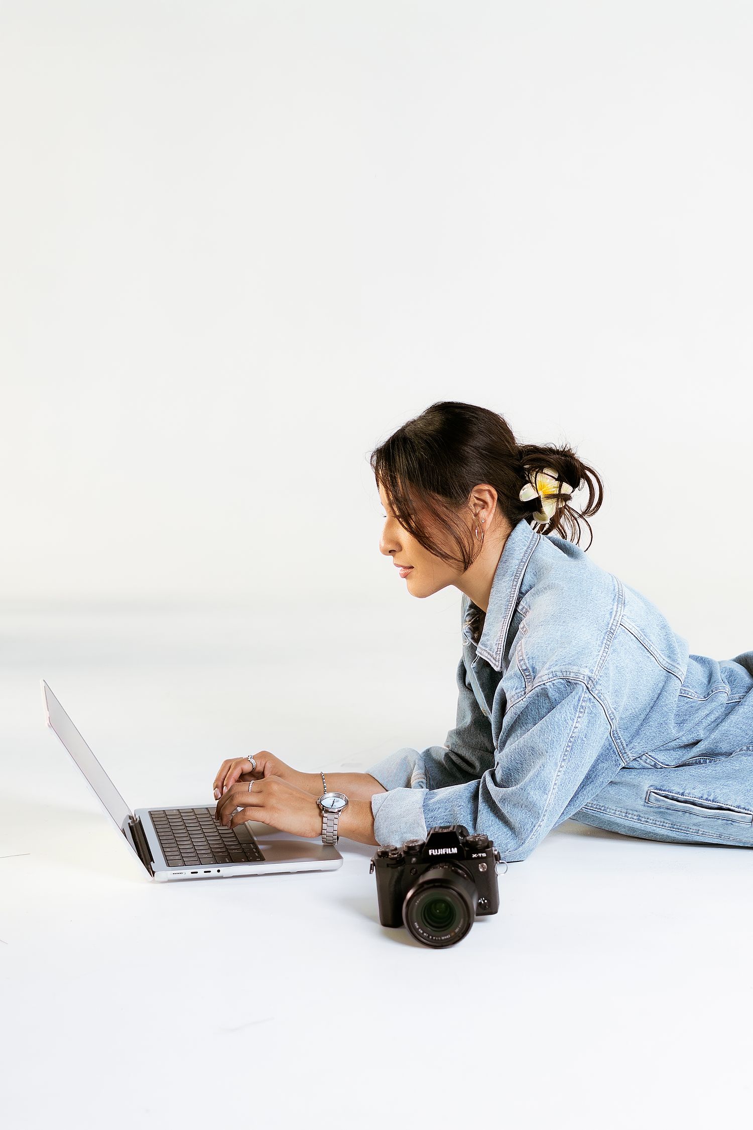 A person in a denim jacket lies on a white surface while working on a laptop and camera in a minimalist setting.