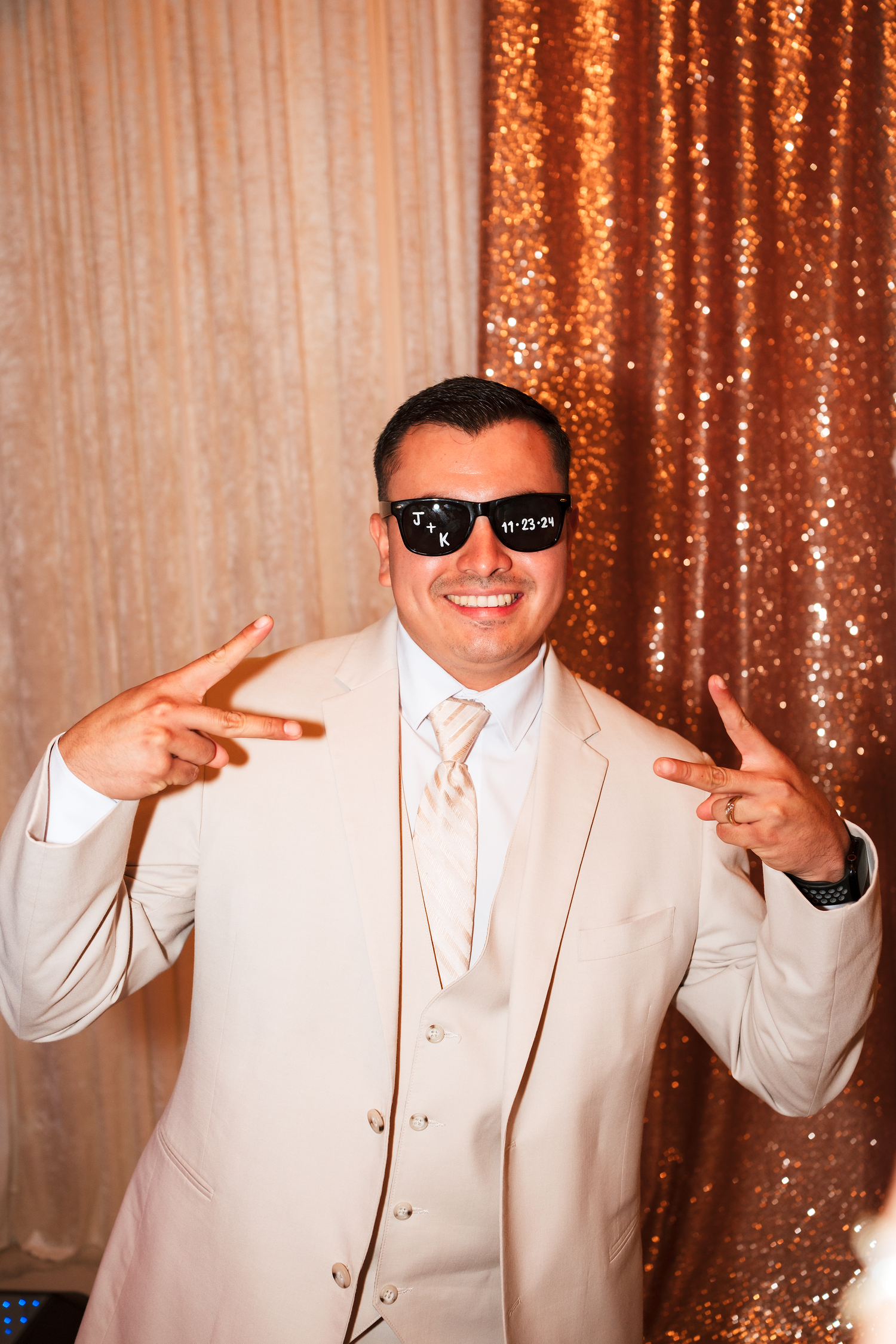 Smiling groom in cream suit poses with sunglasses showing wedding date at reception photo booth.