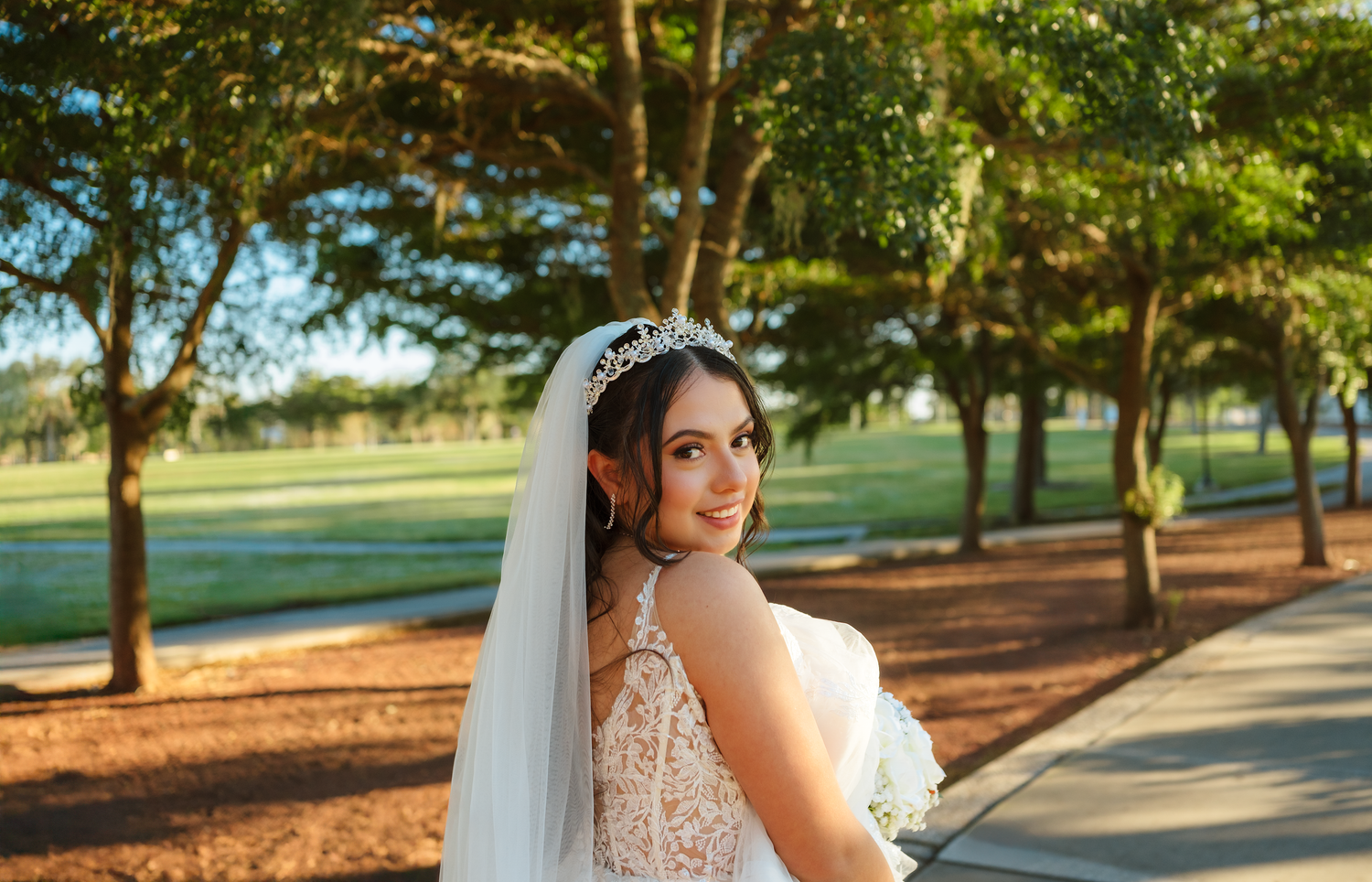 Bride in white lace gown and veil with floral crown smiling over shoulder outdoors near tree-lined path.