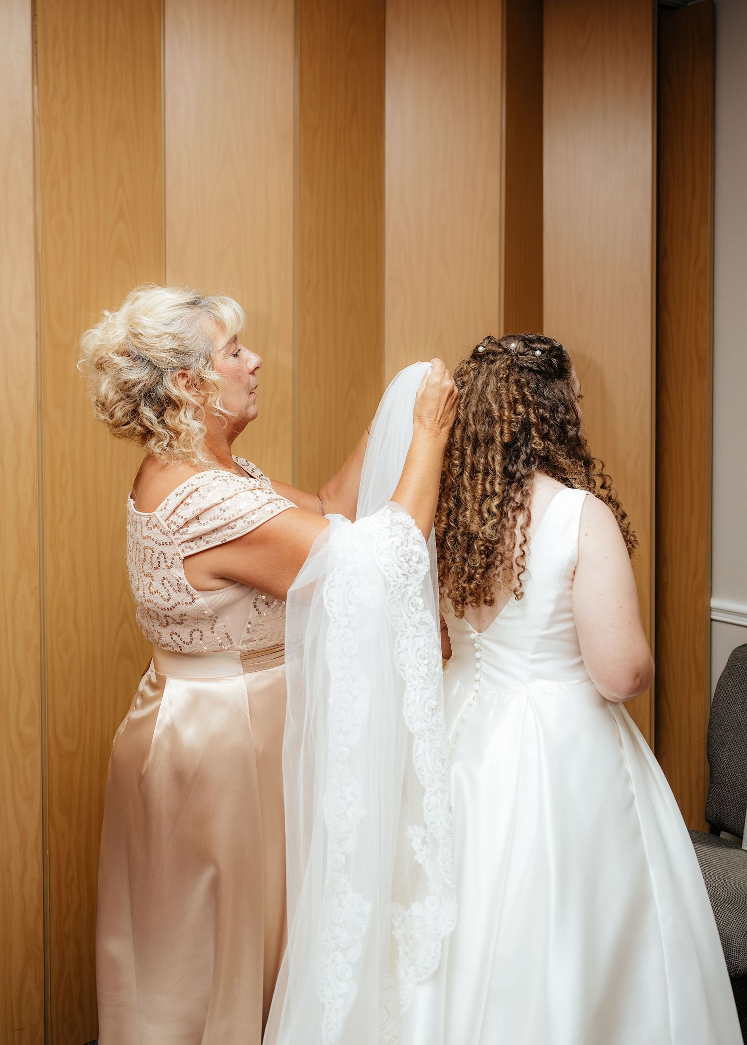 A blonde woman helps a curly-haired bride adjust her white veil before the wedding ceremony.