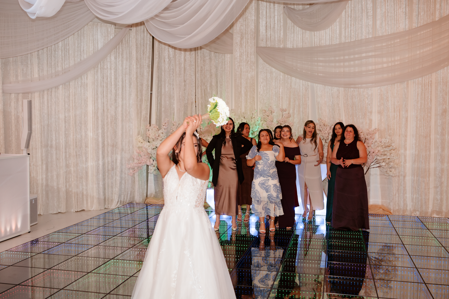 Bride tosses bouquet to excited bridesmaids in elegant white draped wedding reception venue.