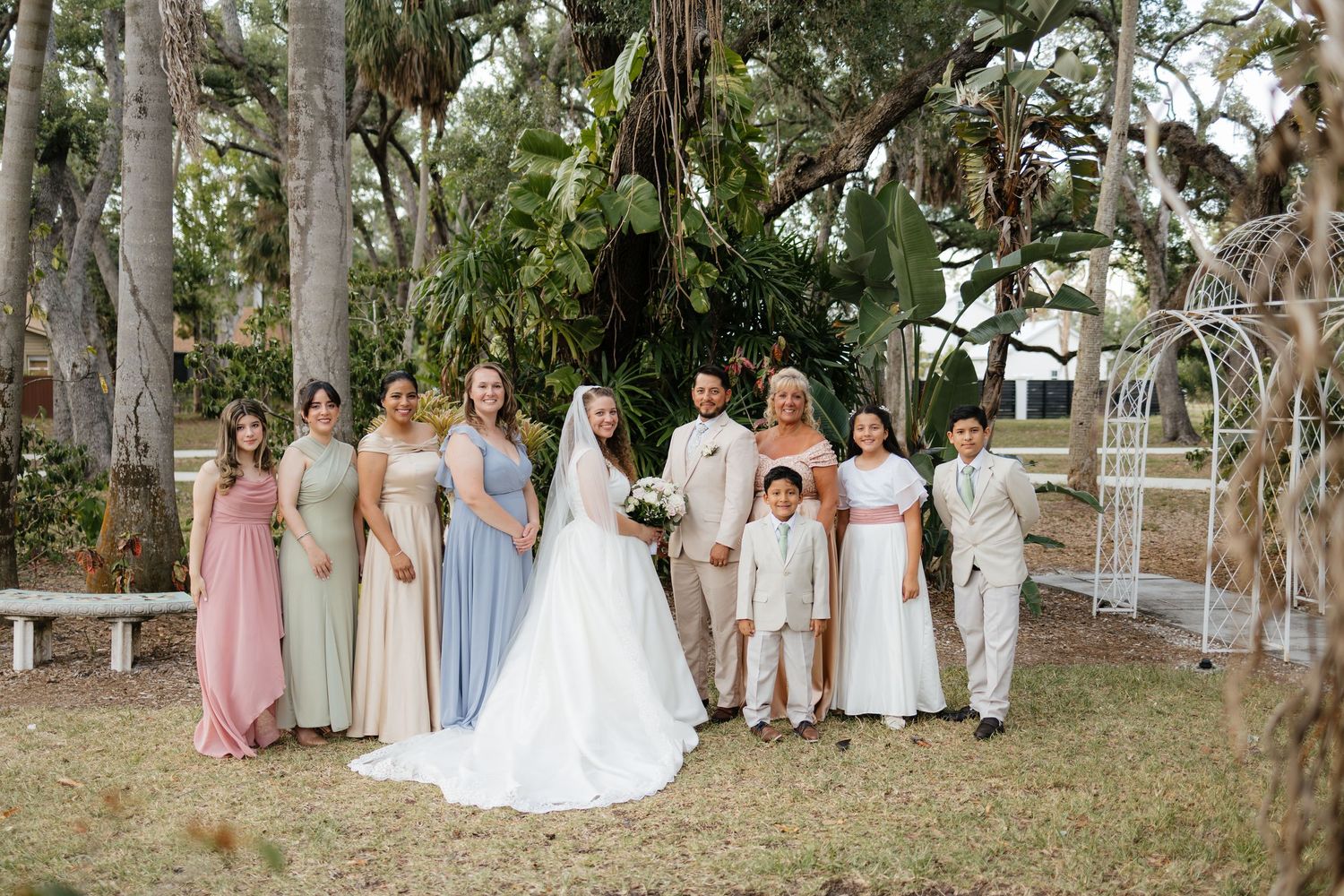 Wedding party poses outdoors under large trees with bride, groom, bridesmaids in pastel gowns, and groomsmen.