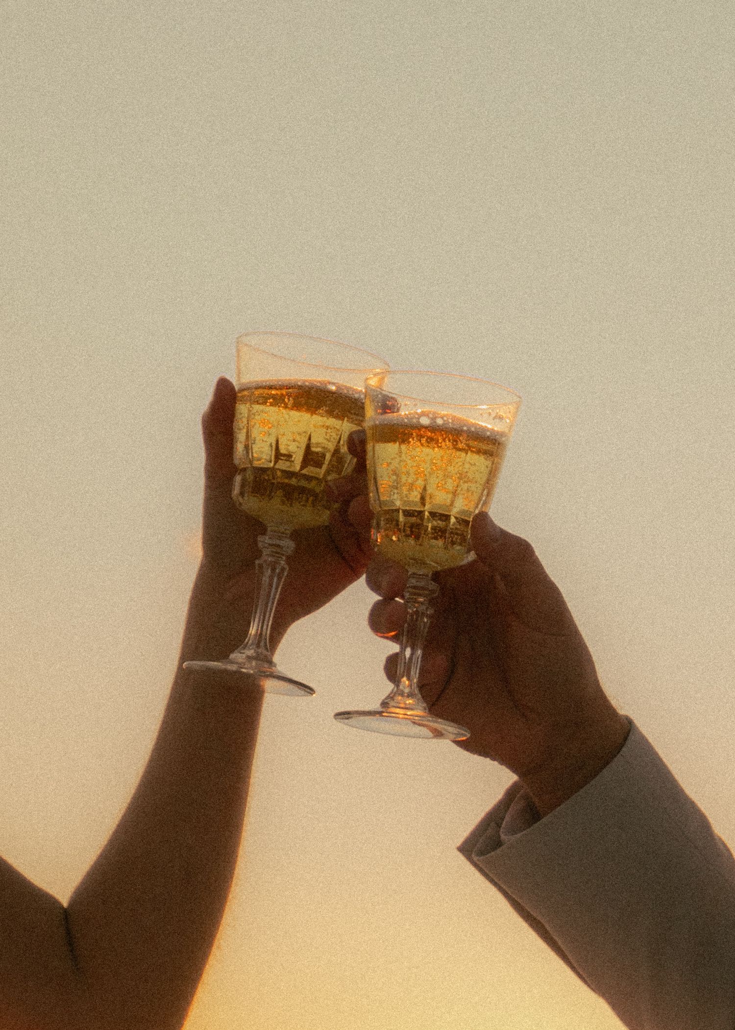 Two hands clinking wine glasses filled with champagne against a warm golden sunset sky.