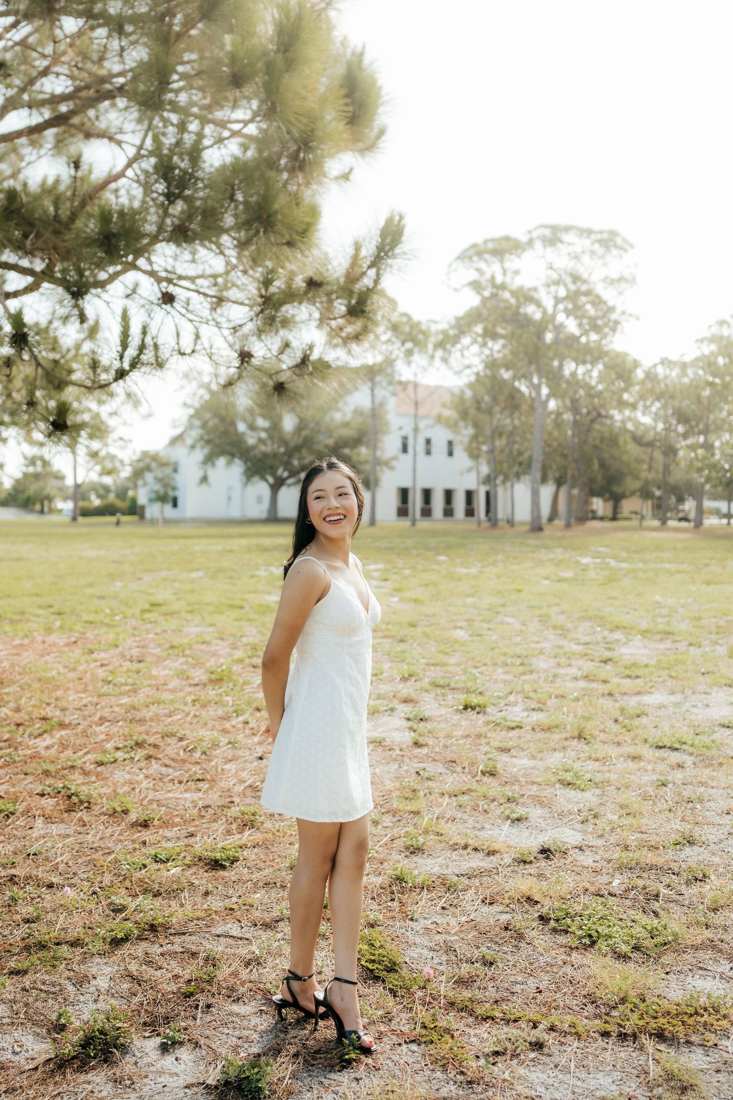 Young Asian woman in white dress and heels smiling outdoors on grassy field with trees and white building background.