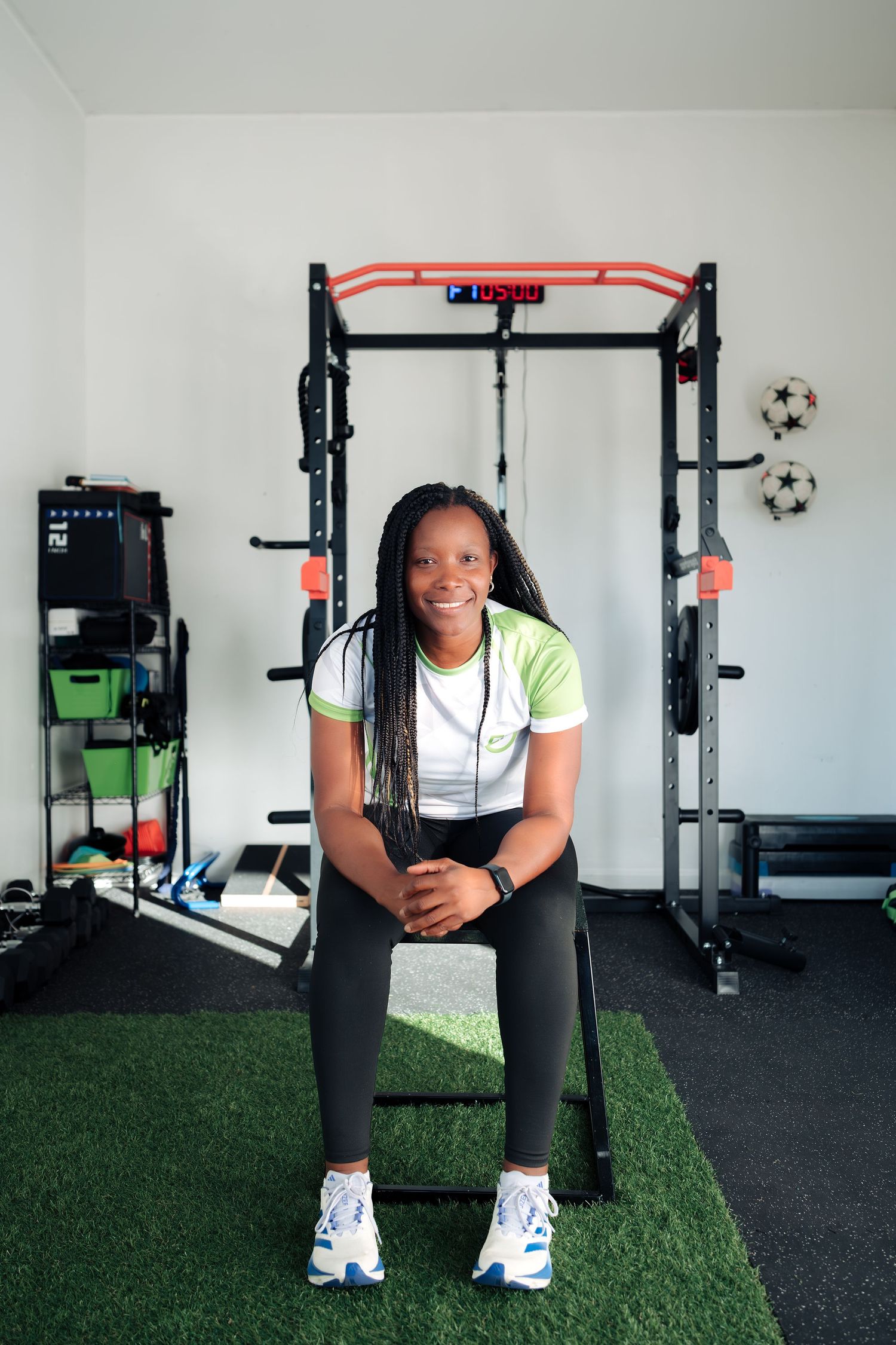 A smiling woman in athletic wear sits in a home gym with a power rack, turf flooring, and fitness equipment.