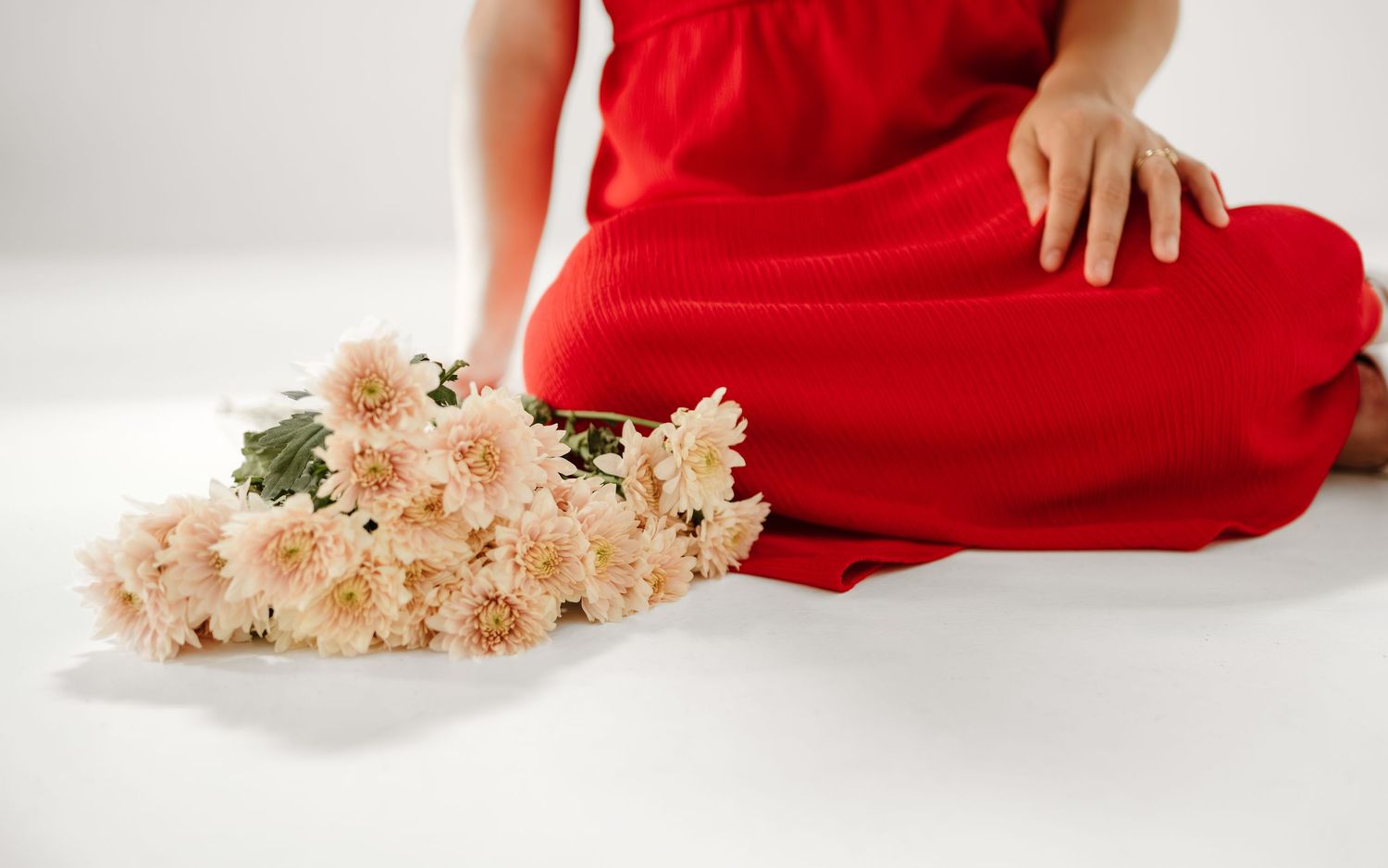 Woman in red dress sitting on white floor with bouquet of soft pink chrysanthemums beside her.