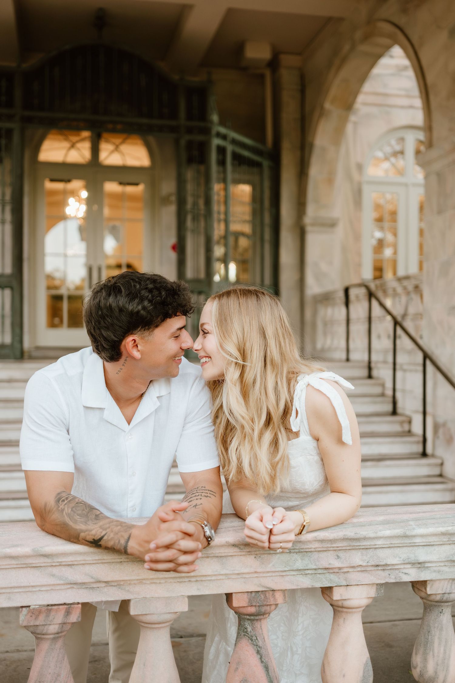 A couple in white outfits smiles at each other while leaning on a stone balustrade outside an elegant building.