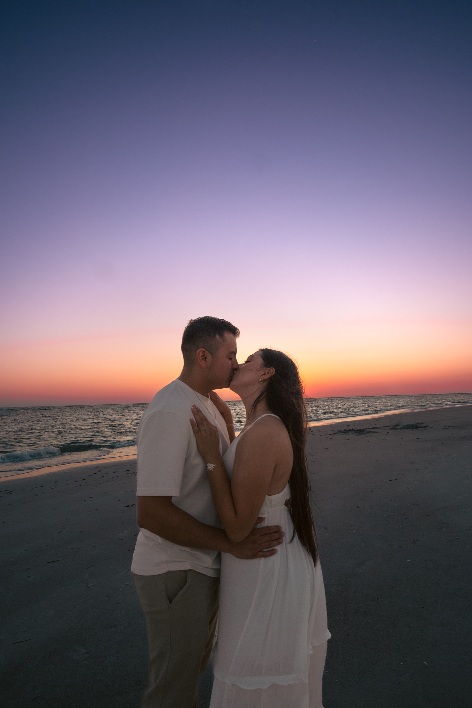Couple sharing a romantic kiss on the beach at sunset with colorful purple and orange sky backdrop.